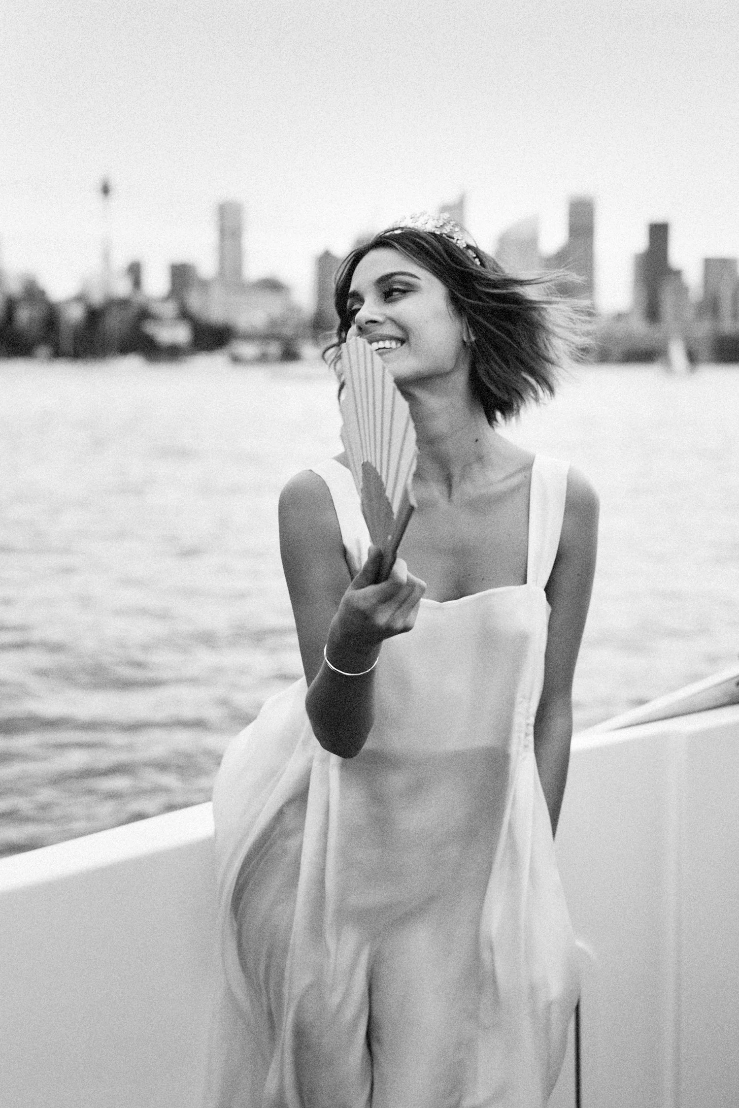 A black and white photo of a smiling woman in a dress holding a fan, standing near water with a city skyline in the background.