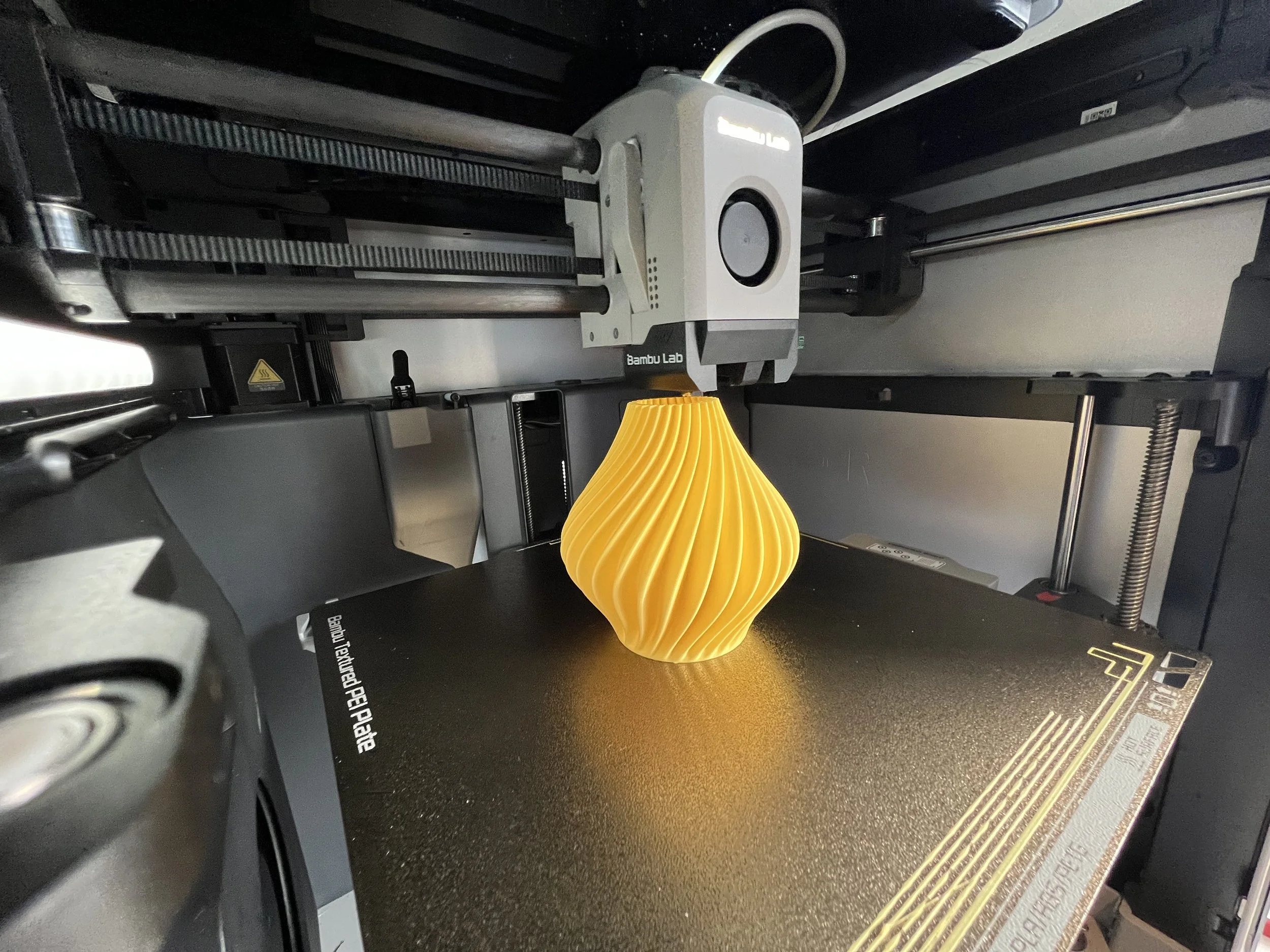 A 3D printed yellow vase with a spiral design on a 3D printer's build platform.