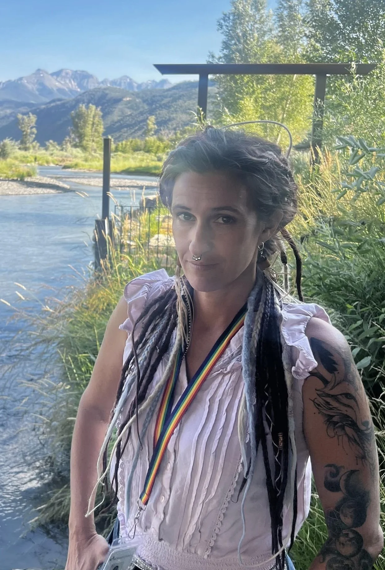 A woman with long dreadlocks, tattoos, and a septum piercing standing outdoors by a river, with mountains in the background, wearing a rainbow-colored lanyard.