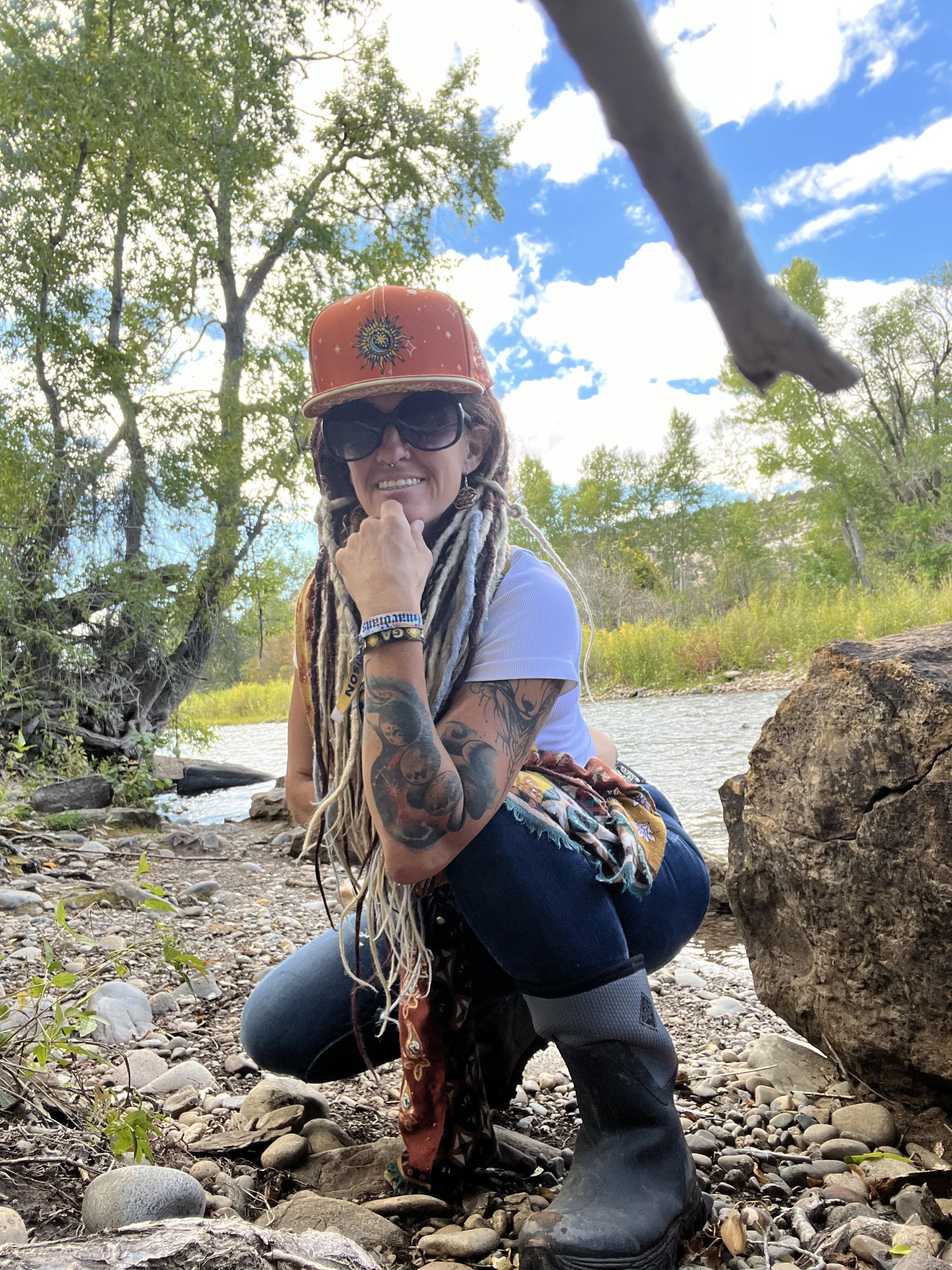 A woman with long dreadlocks, tattoos, large sunglasses, and a red cap kneeling near a riverbank with rocks and trees, smiling at the camera.