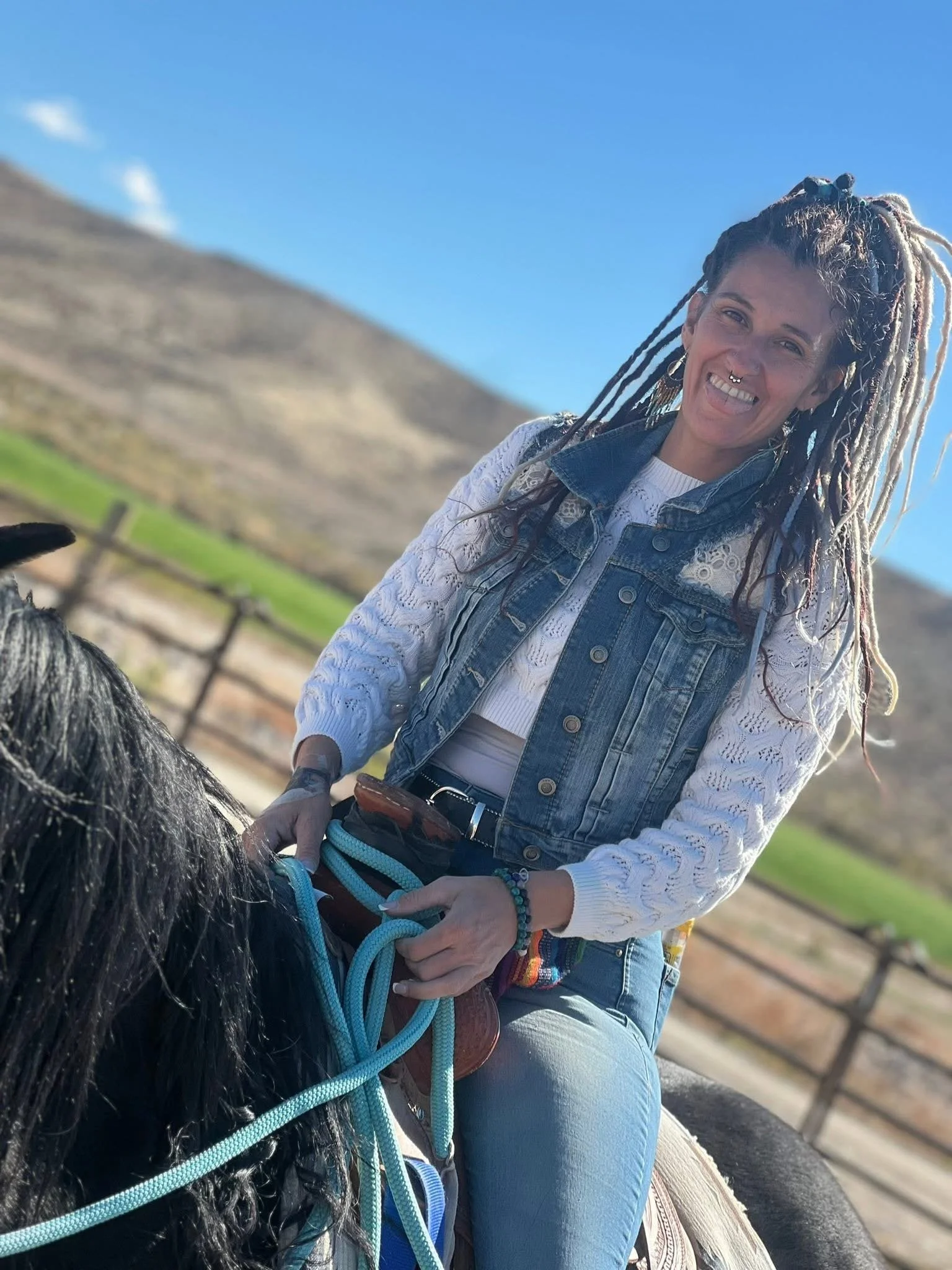 A woman with dreadlocks and a septum piercing riding a horse outdoors with a mountain and blue sky in the background.