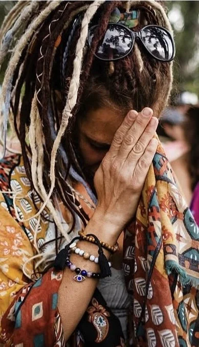 A woman with dreadlocks and sunglasses on her head, covering her face with her hand, wearing colorful clothing and bracelets.