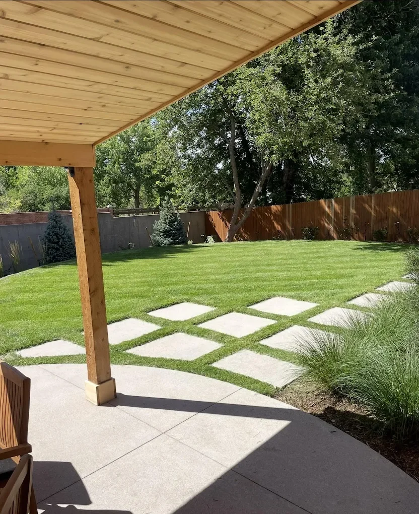 View of a backyard with a covered patio, green grass lawn, concrete stepping stones, and trees and shrubs along the fence line.