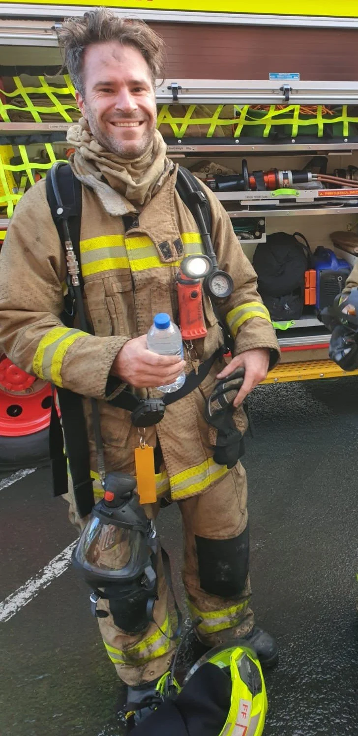 Dr. Jean Renouf, as a firefighter in full gear, standing in front of a fire truck with equipment mounted on the side.