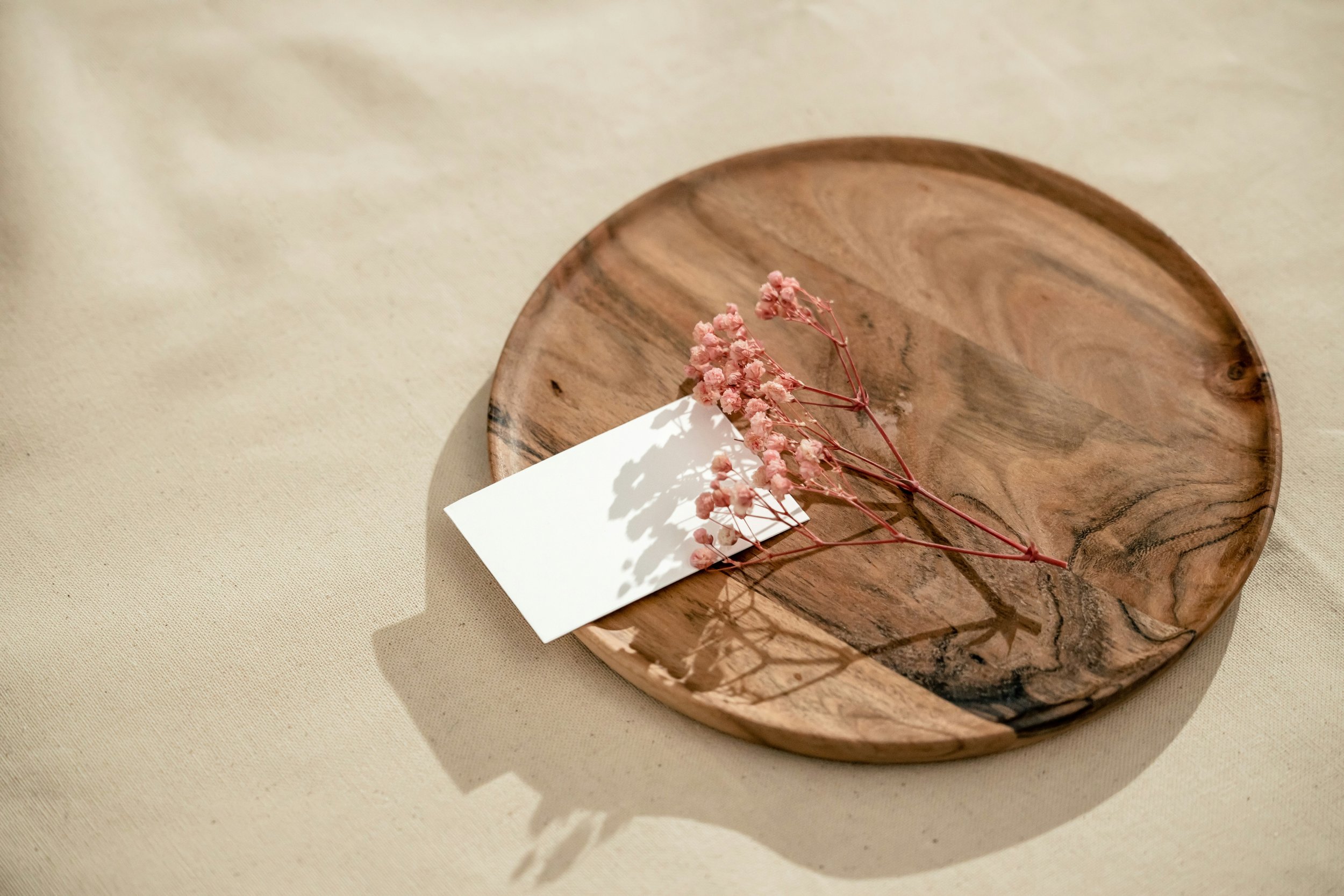 A wooden plate with pink dried flowers and a blank white note card on a beige fabric surface.