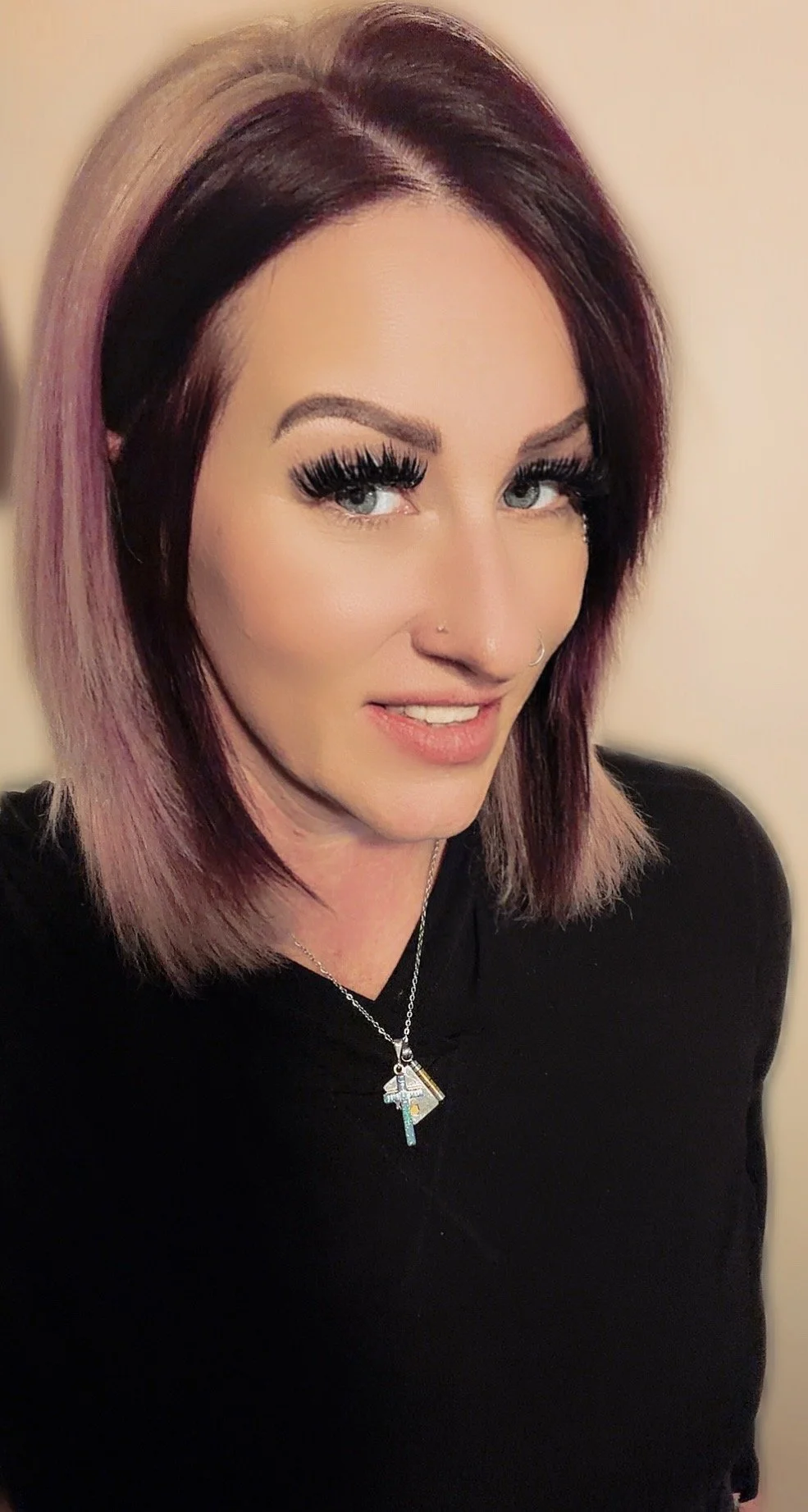 A woman with shoulder-length purple and brown hair, wearing makeup with dramatic eyelashes, a nose piercing, and a necklace with cross pendants, smiling and looking at the camera.