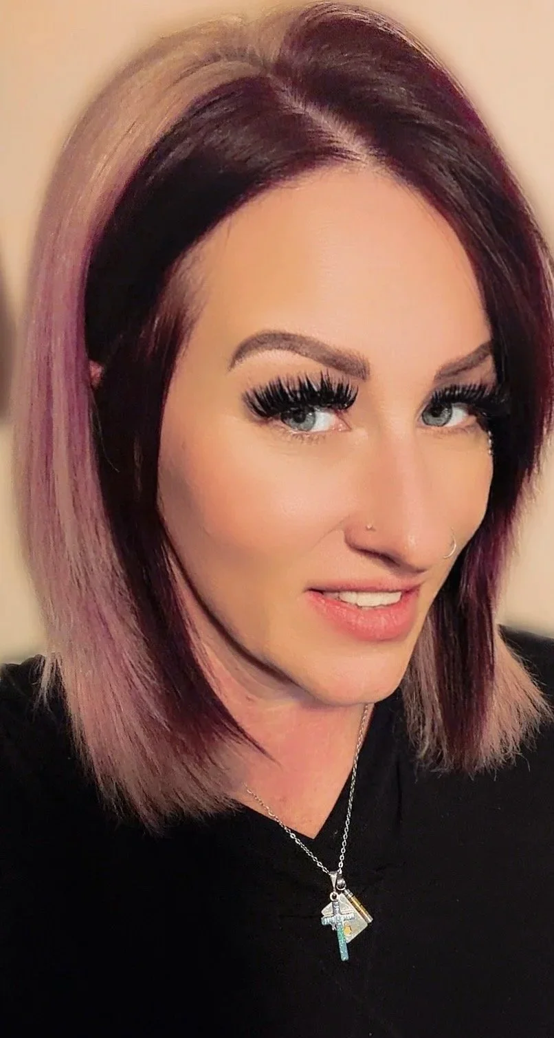 A woman with shoulder-length hair styled in a bob, with dark roots and purple-pink highlights, wearing heavy makeup, long false eyelashes, and a nose ring, smiling slightly, wearing a black top and a necklace with a colorful pendant.