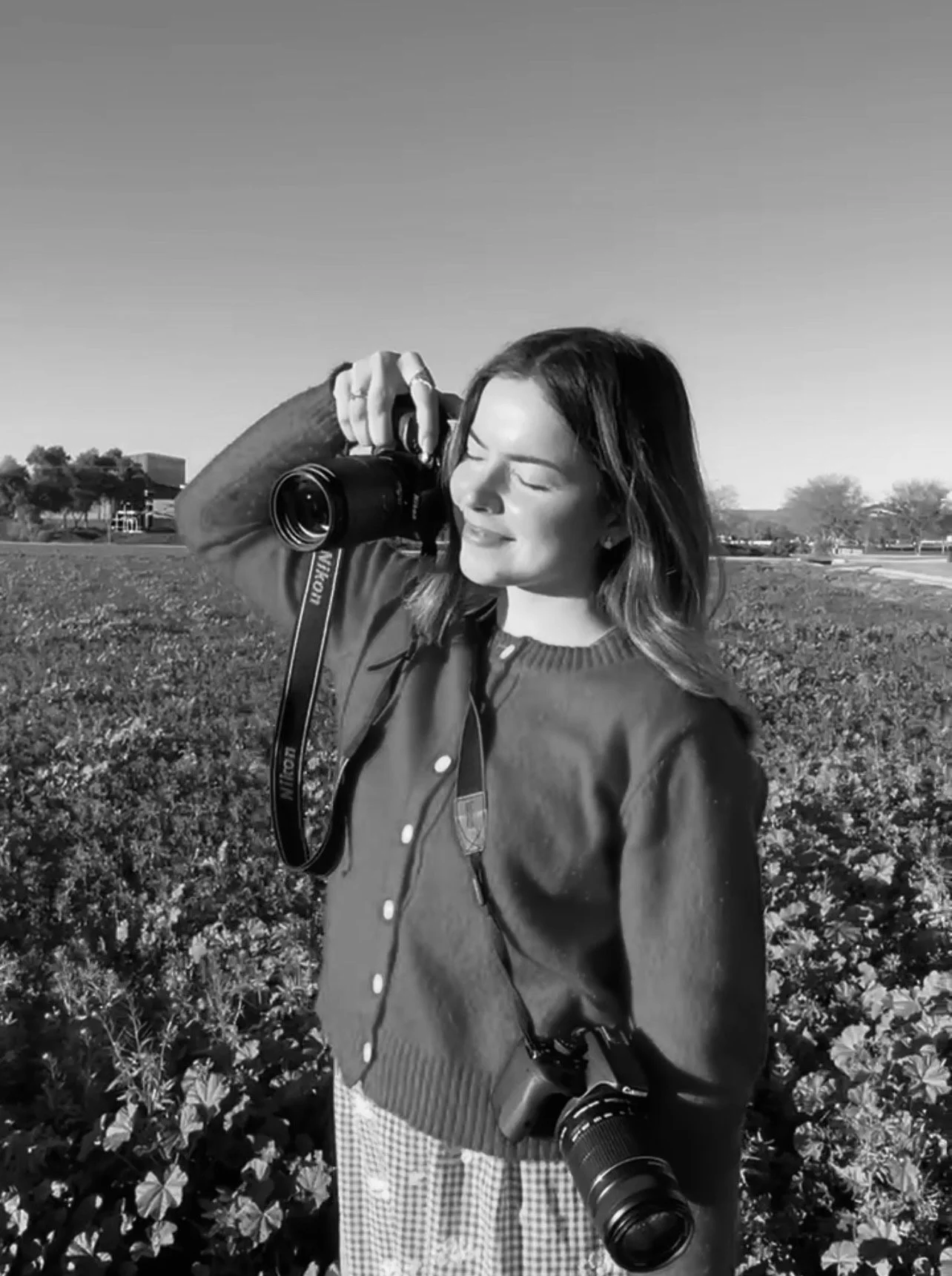 A woman with shoulder-length hair, wearing a cardigan and checkered skirt, is smiling with eyes closed, taking a photograph outdoors in a field of flowers, with a second camera hanging from her neck.