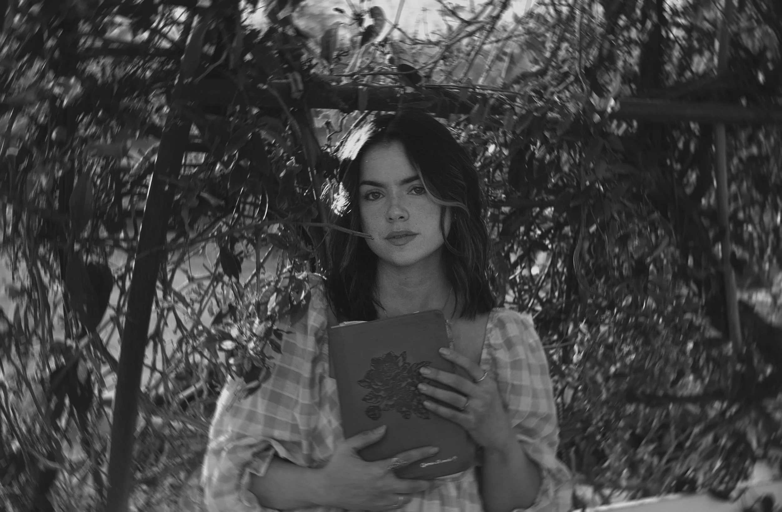 A woman with dark hair, wearing a checkered dress, holding a book, standing among dense foliage in black and white.
