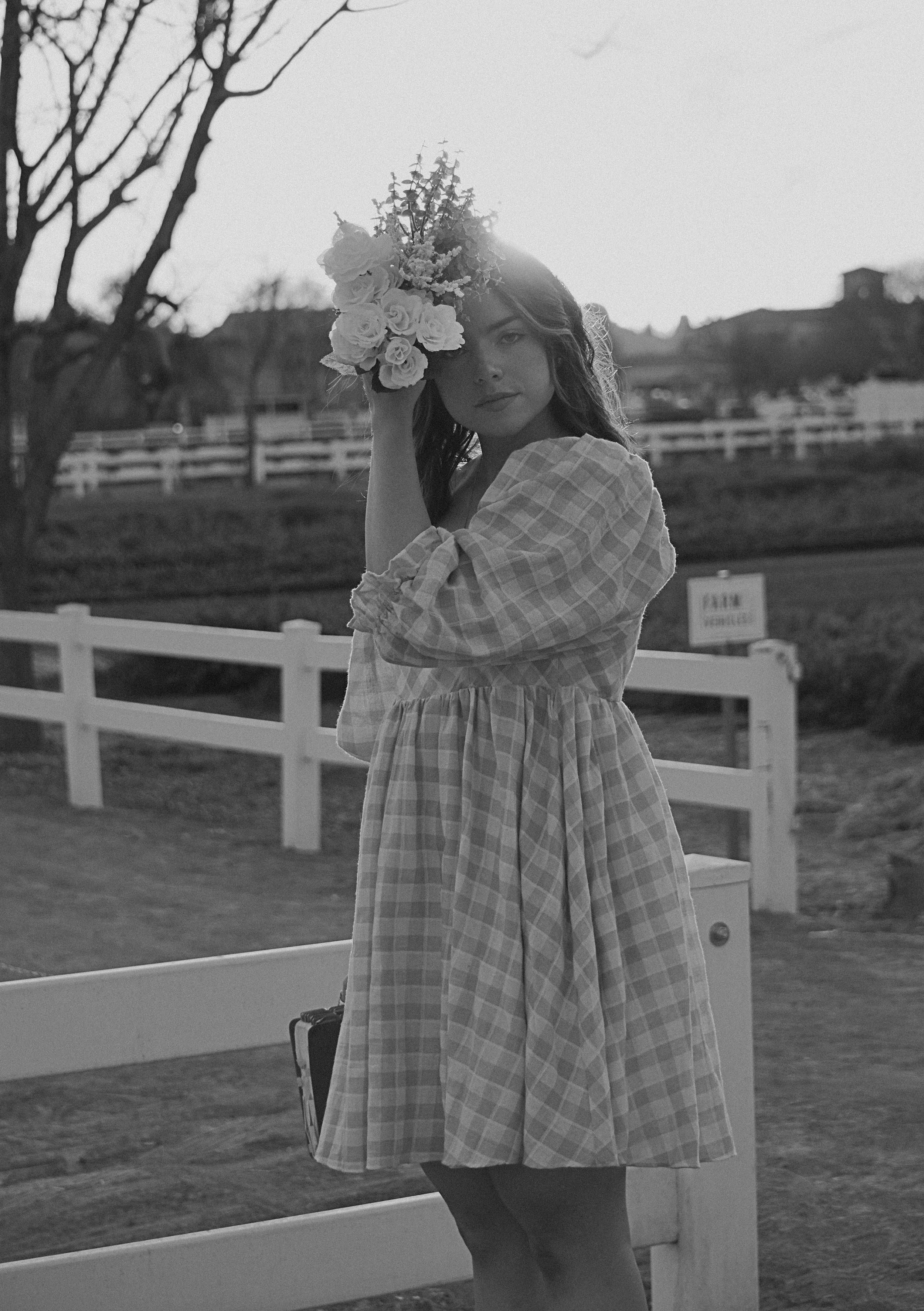 A woman in a checkered dress holding a bouquet of flowers on her head, standing outdoors near a white fence in a rural setting.