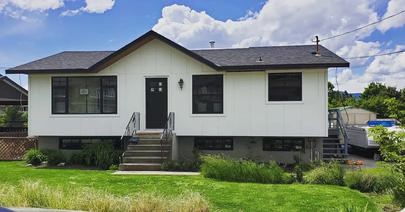 A white house with black trim windows and black stairs leading to the front door. The house has a sloped roof and is elevated with a grassy yard in front and a cloudy sky above.