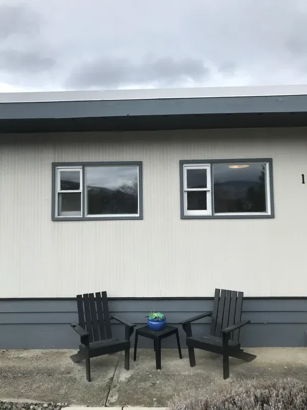 Two black Adirondack chairs with a small black table between them, with a blue pot containing a plant, in front of a beige house with two windows and a cloudy sky above.