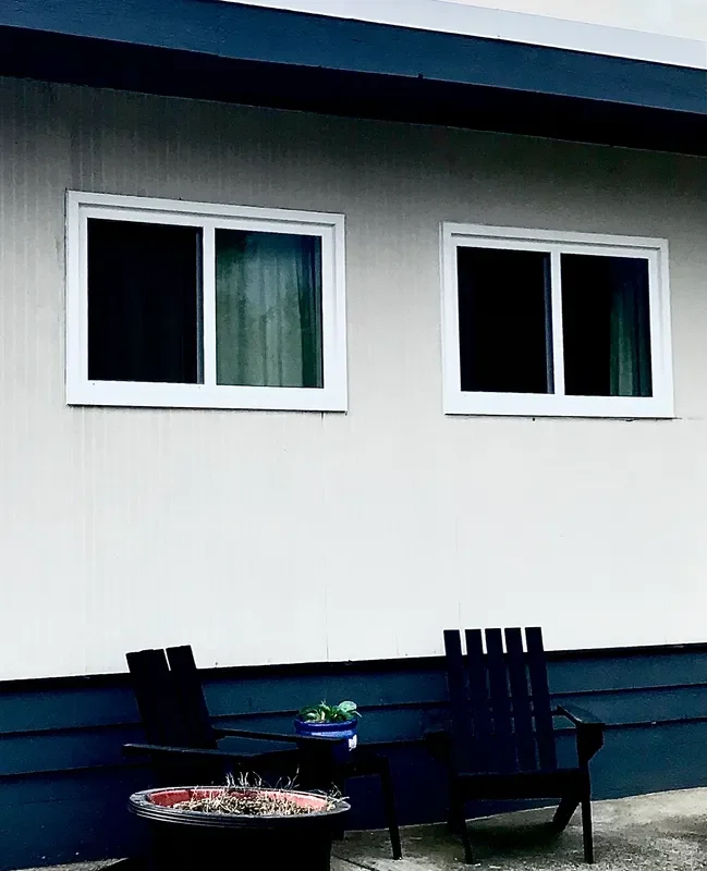 Close-up of a house exterior with two white-framed windows, dark siding, and a small outdoor seating area with two black chairs and a blue pot with plants.