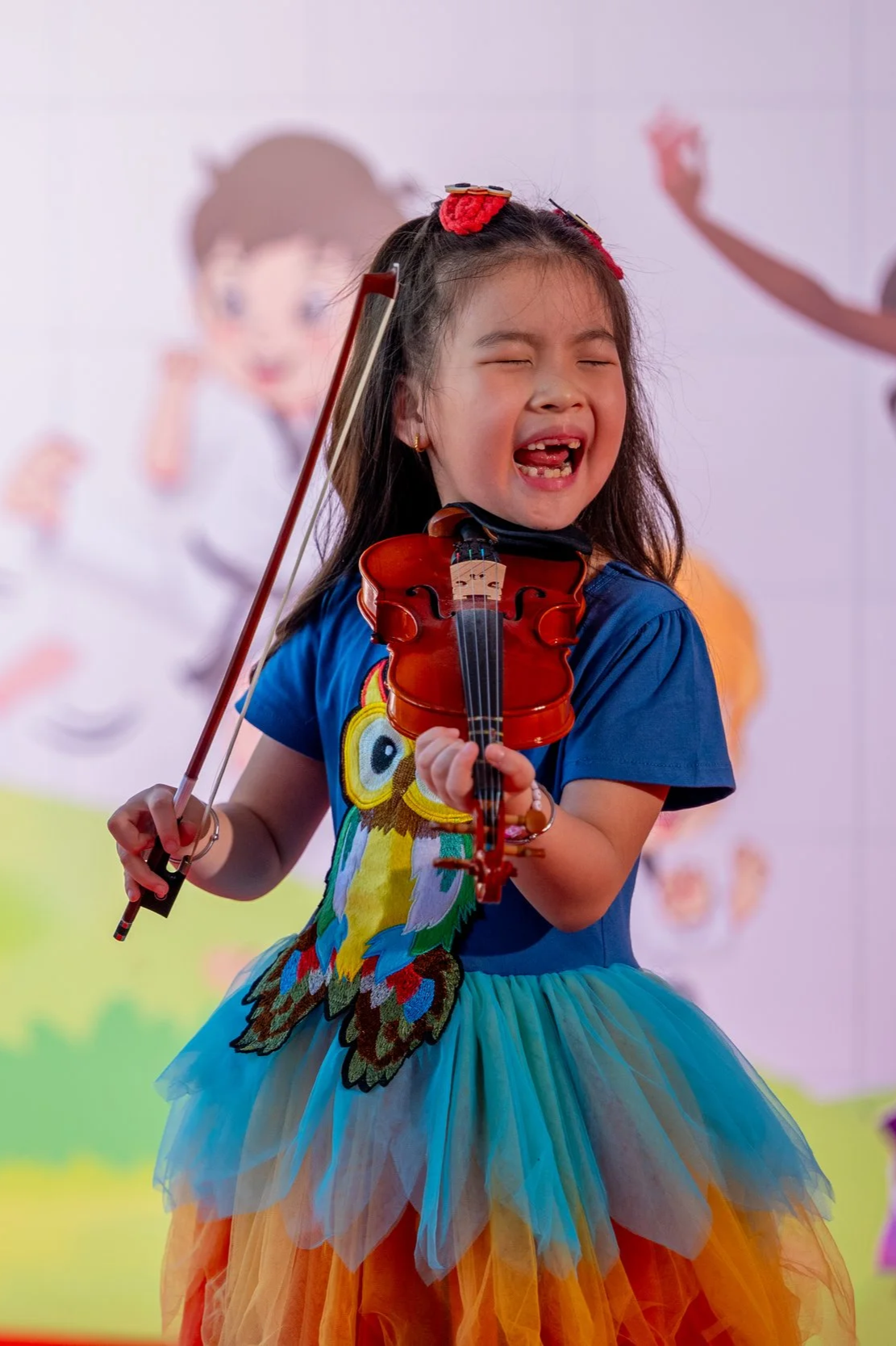 A young girl with long dark hair playing a violin and smiling with her eyes closed. She is wearing a blue top with an owl design on it and a colorful tutu skirt. The background features a cartoon character on a white wall.