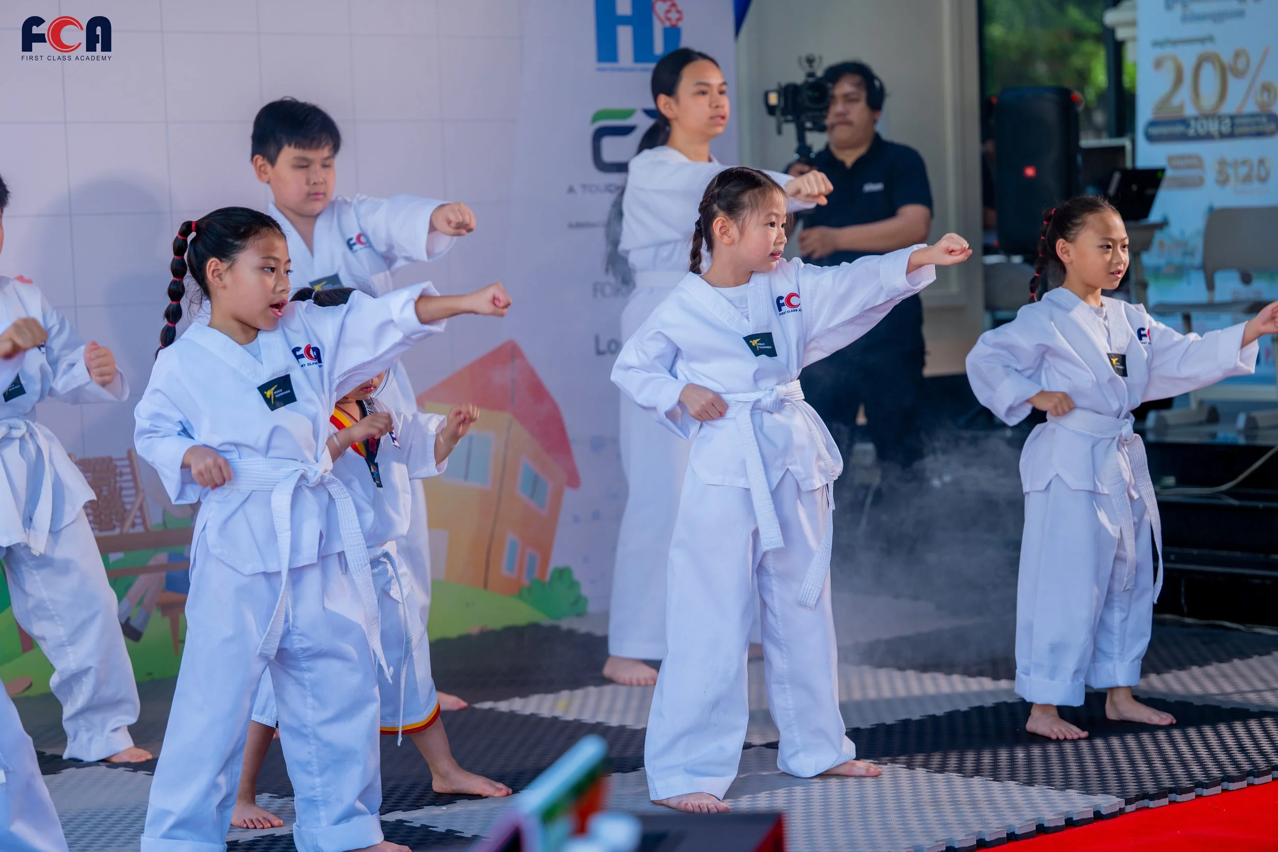 Children practicing martial arts on a stage, wearing white uniforms with belts, with a backdrop featuring logos and a camera person recording the event.