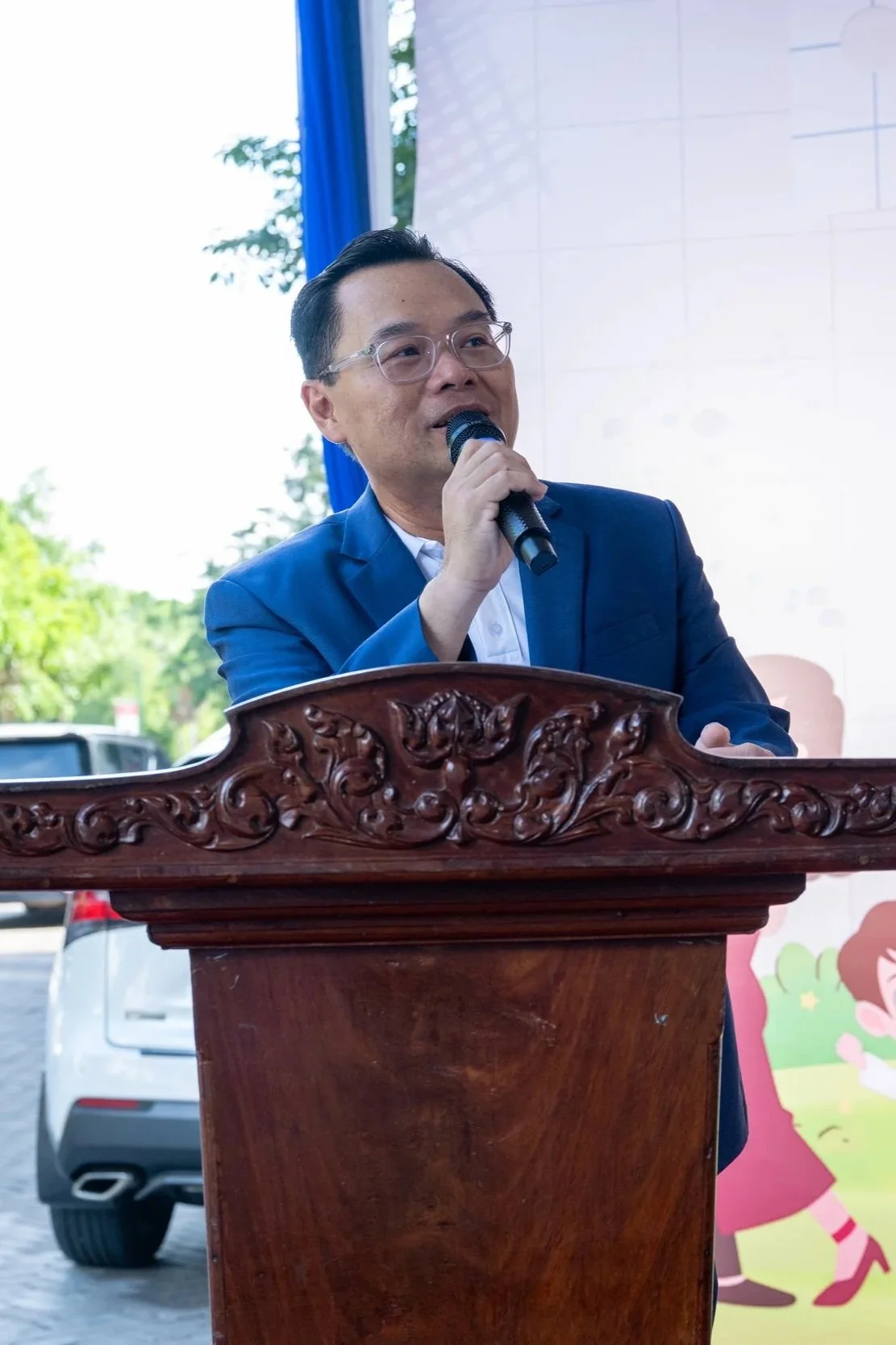 A man in a blue suit and glasses speaking into a microphone at a podium with carved wood details during an outdoor event.