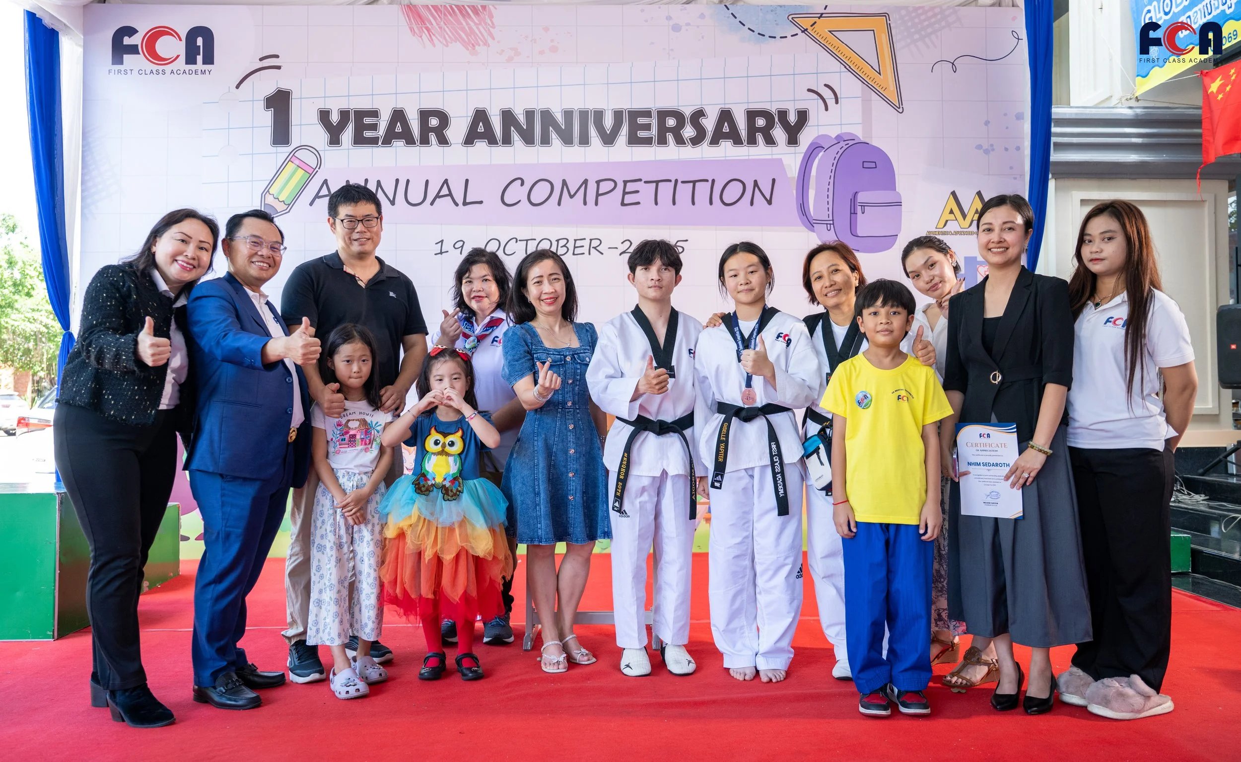 Group of people, including children and adults, standing on a stage with a banner in the background that reads '1 Year Anniversary Annual Competition, 19 October 2023'. They are smiling, some giving thumbs-up signs. The children include a girl in a colorful owl costume and a boy in a yellow shirt. The adults are dressed in casual and business attire. The event appears to celebrate the first anniversary of an educational institution or program.