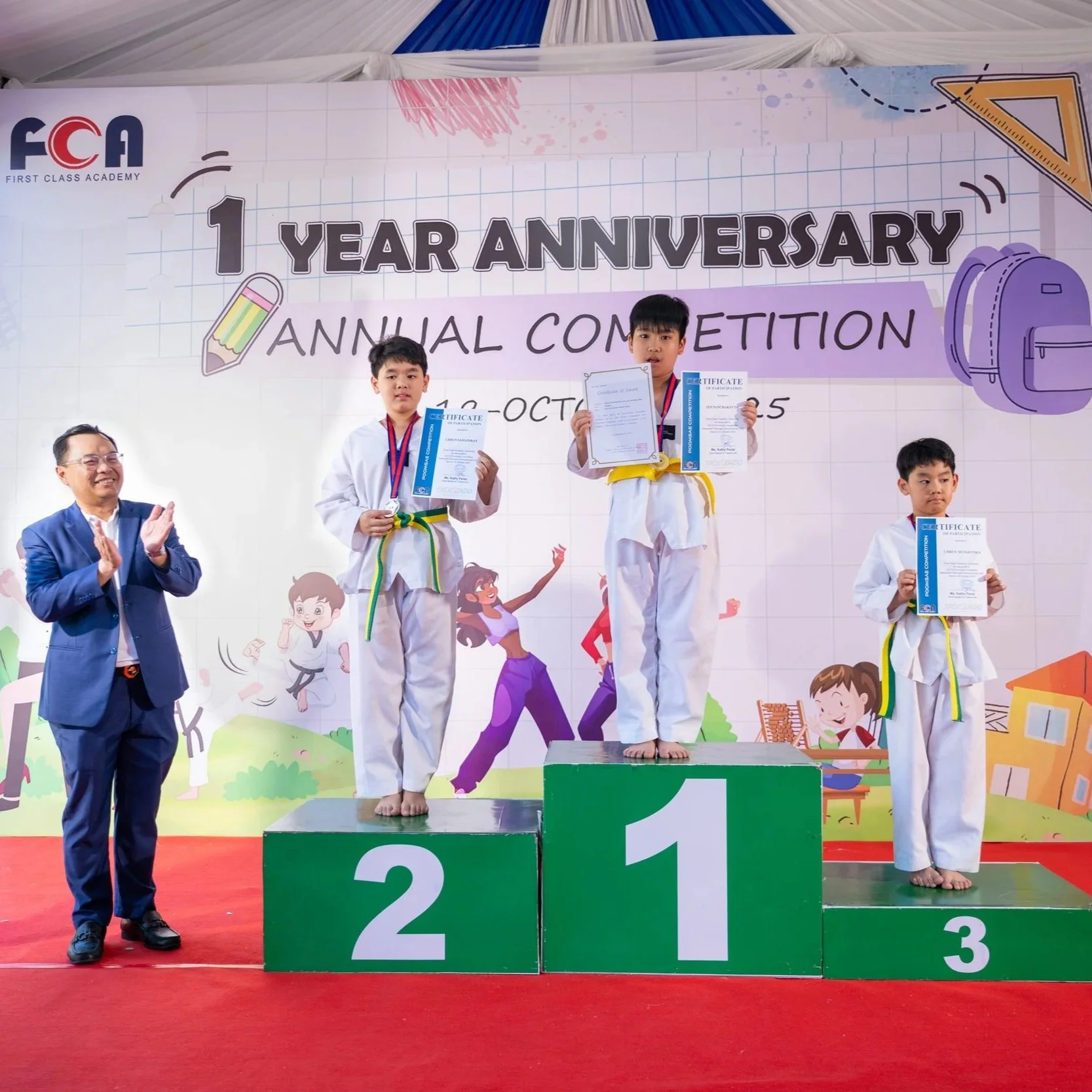 Children in martial arts uniforms on an awards podium holding certificates during a 1st anniversary competition at First Class Academy, with an adult applauding beside them.