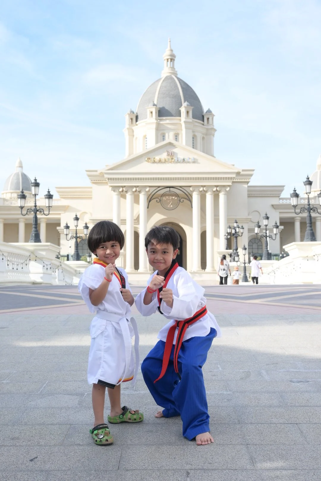 Two children practicing martial arts in front of a grand, white building with classical architecture and a large dome, under clear blue skies.