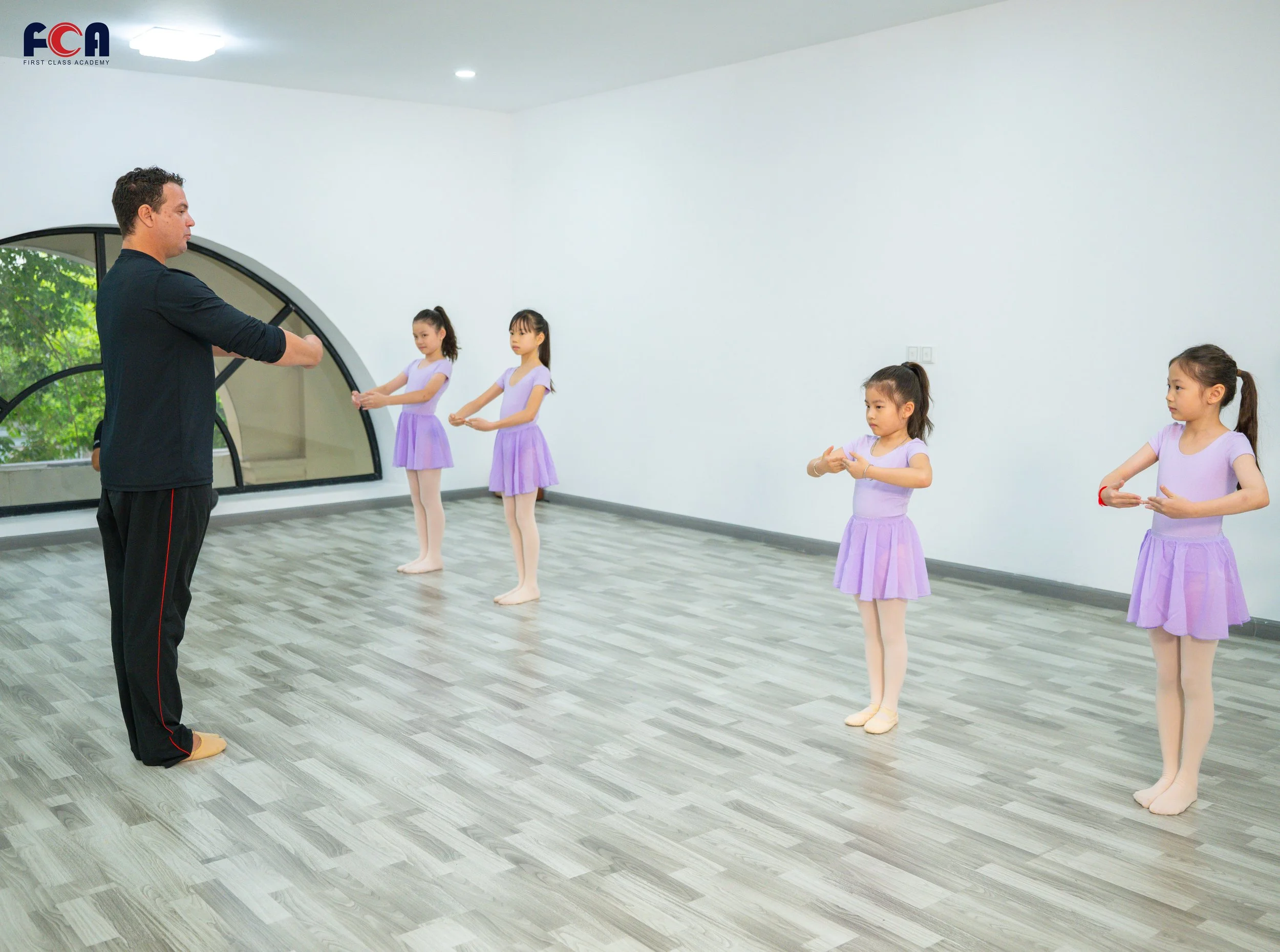 A ballet teacher instructing four young girls dressed in purple leotards and tutus practicing ballet in a dance studio with light gray wood floors and large windows.