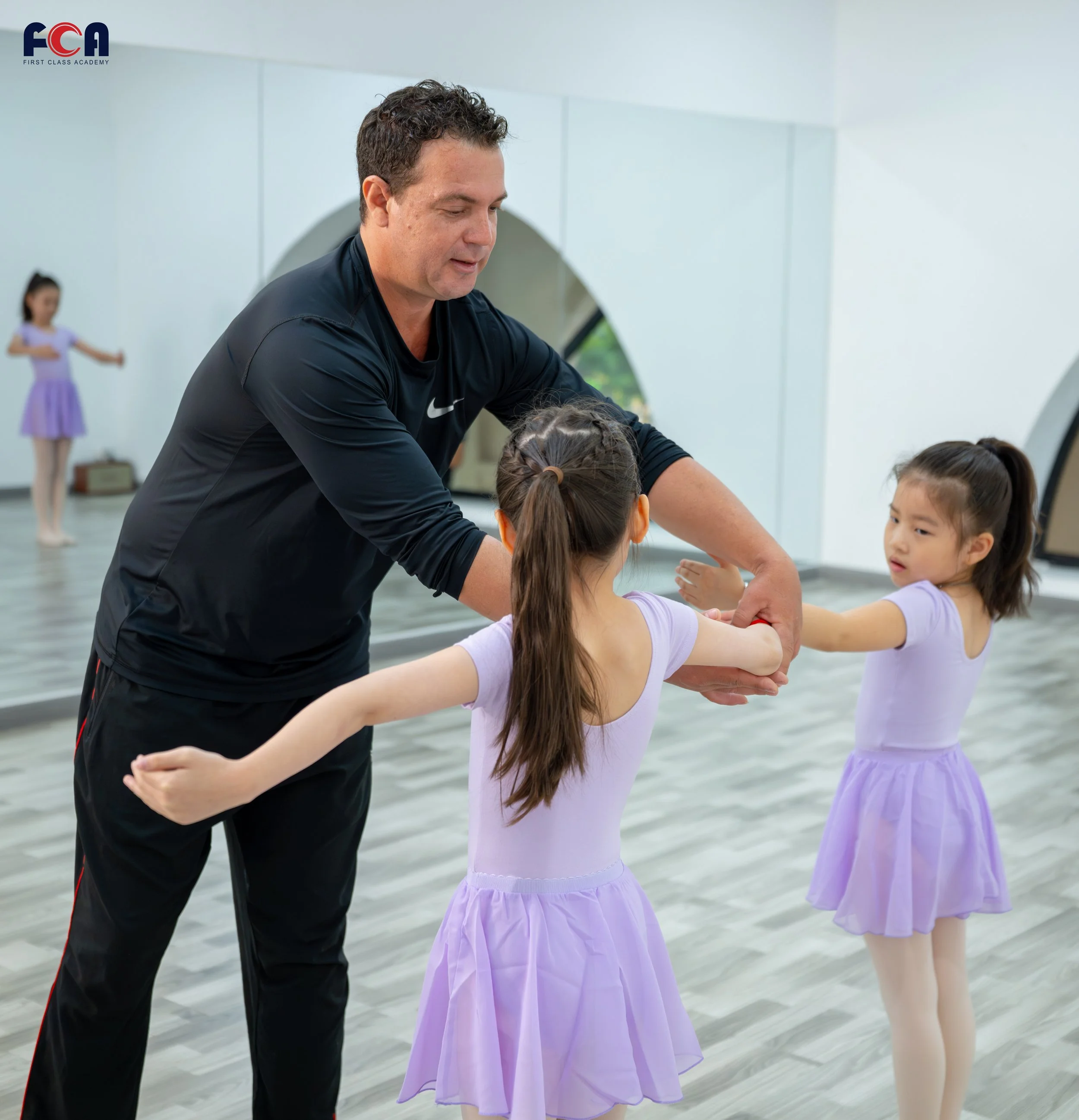 A male ballet instructor in black clothing teaching two young girls in lavender tutus in a dance studio with a mirror and wooden floor.