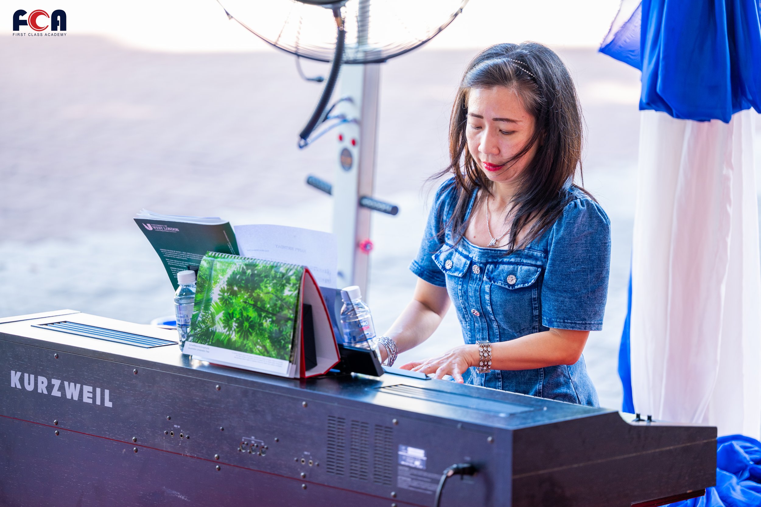 A woman in a denim dress playing a Kurzweil digital piano outdoors with ocean in the background, next to a fan and colorful fabrics.