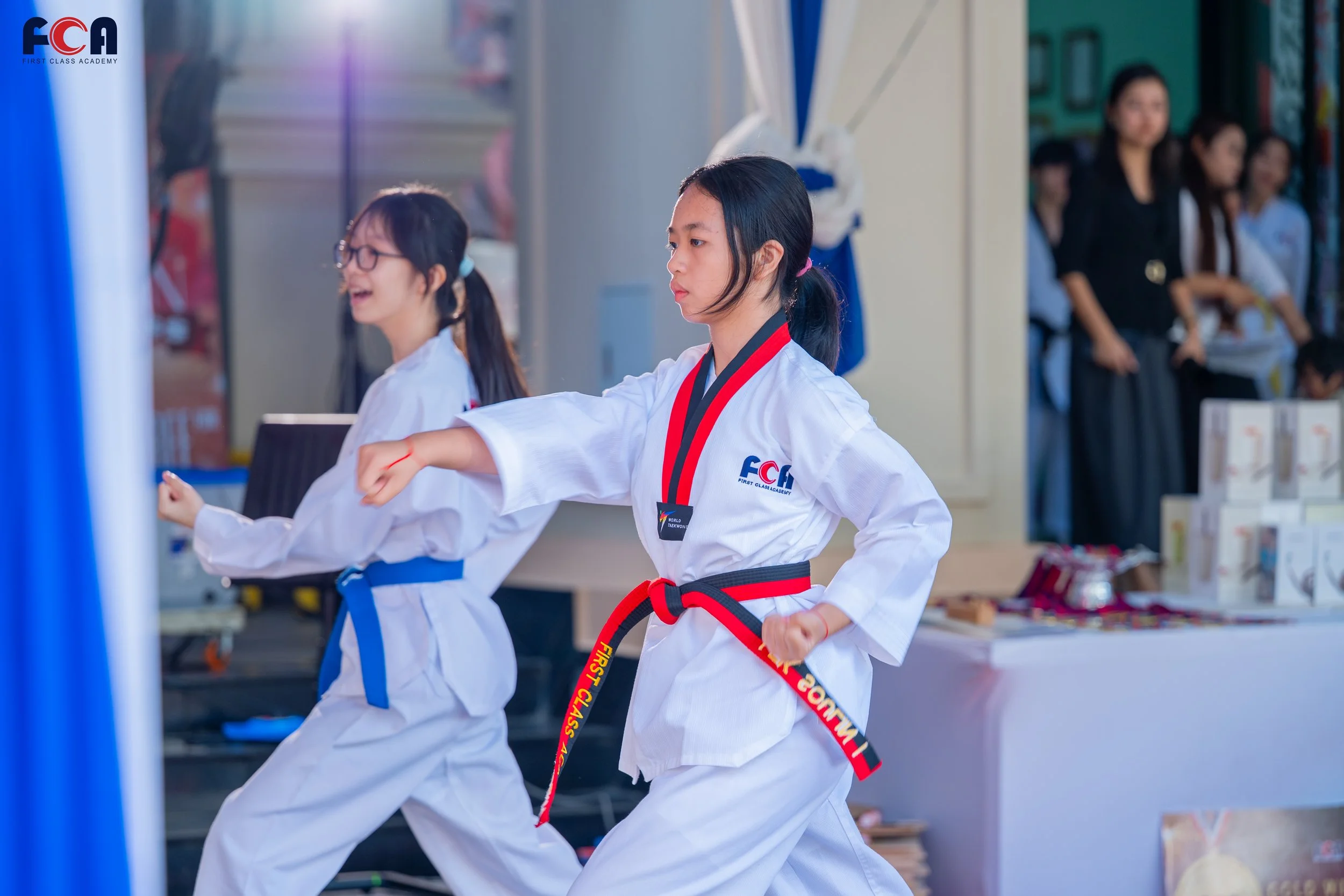 Two young girls practice martial arts in uniforms with colored belts. One girl with a black belt stands in focus, and the other with a blue belt is smiling and slightly blurred. They appear to be inside a school or gym, with onlookers and items displayed in the background.