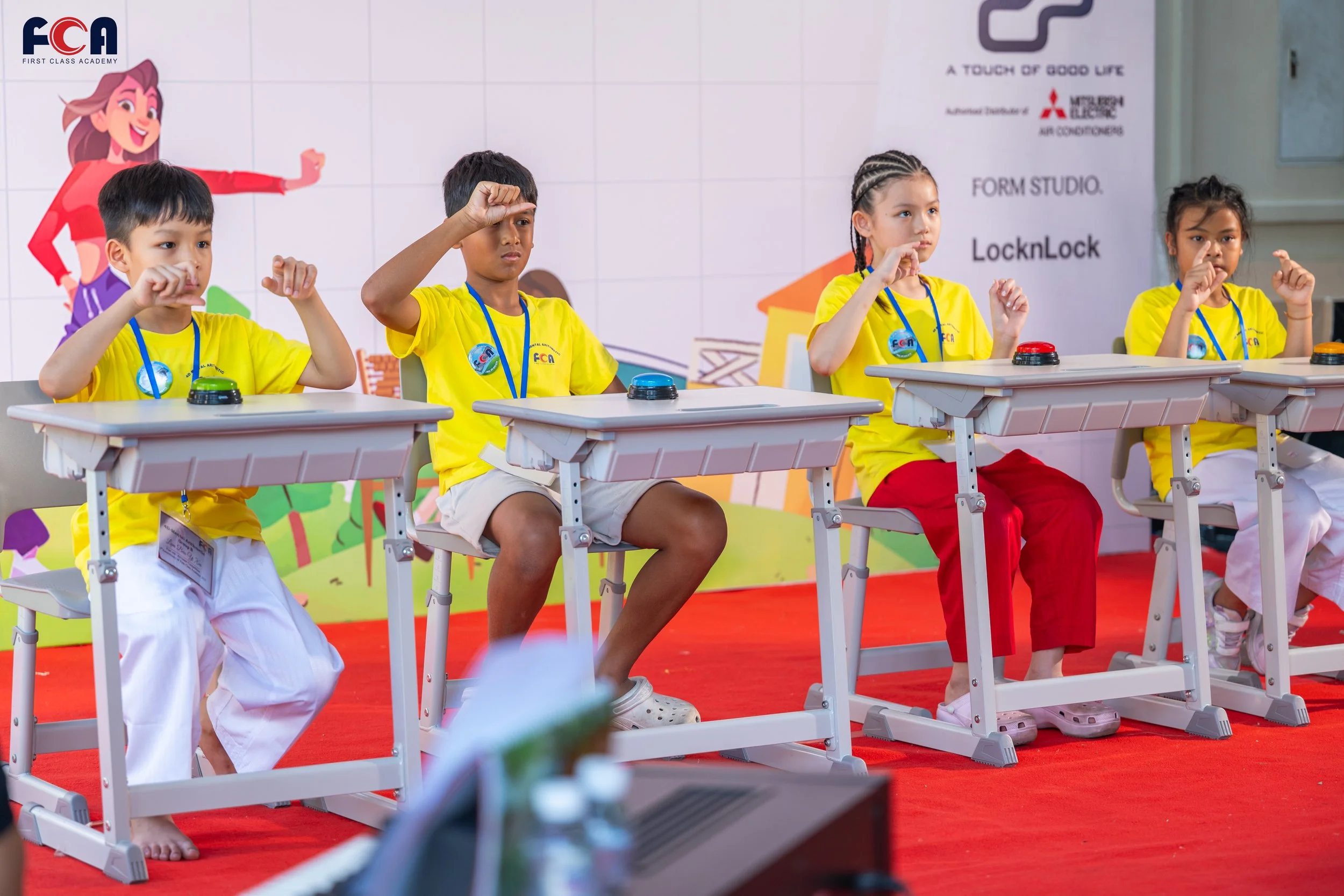 Four children sitting at desks participating in a quiz or game, each with a buzzer in front of them, on a stage with a colorful backdrop featuring cartoon characters and text related to a children's event.