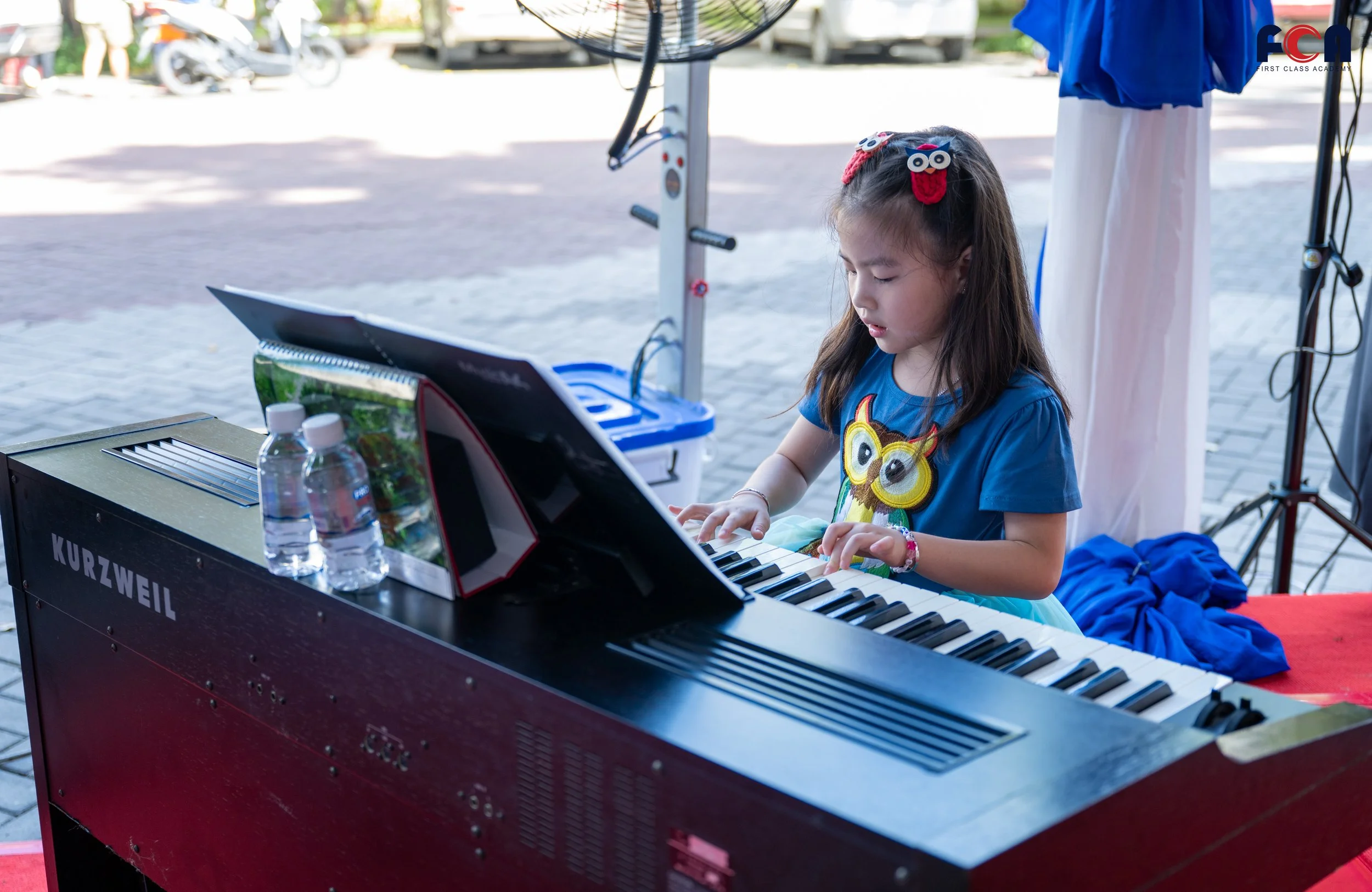 A young girl wearing a blue shirt with an owl design, playing a Kurzweil electric keyboard outdoors. She has long brown hair with owl-shaped hair clips. There are two water bottles on the keyboard and a transparent plastic container with a plant inside.