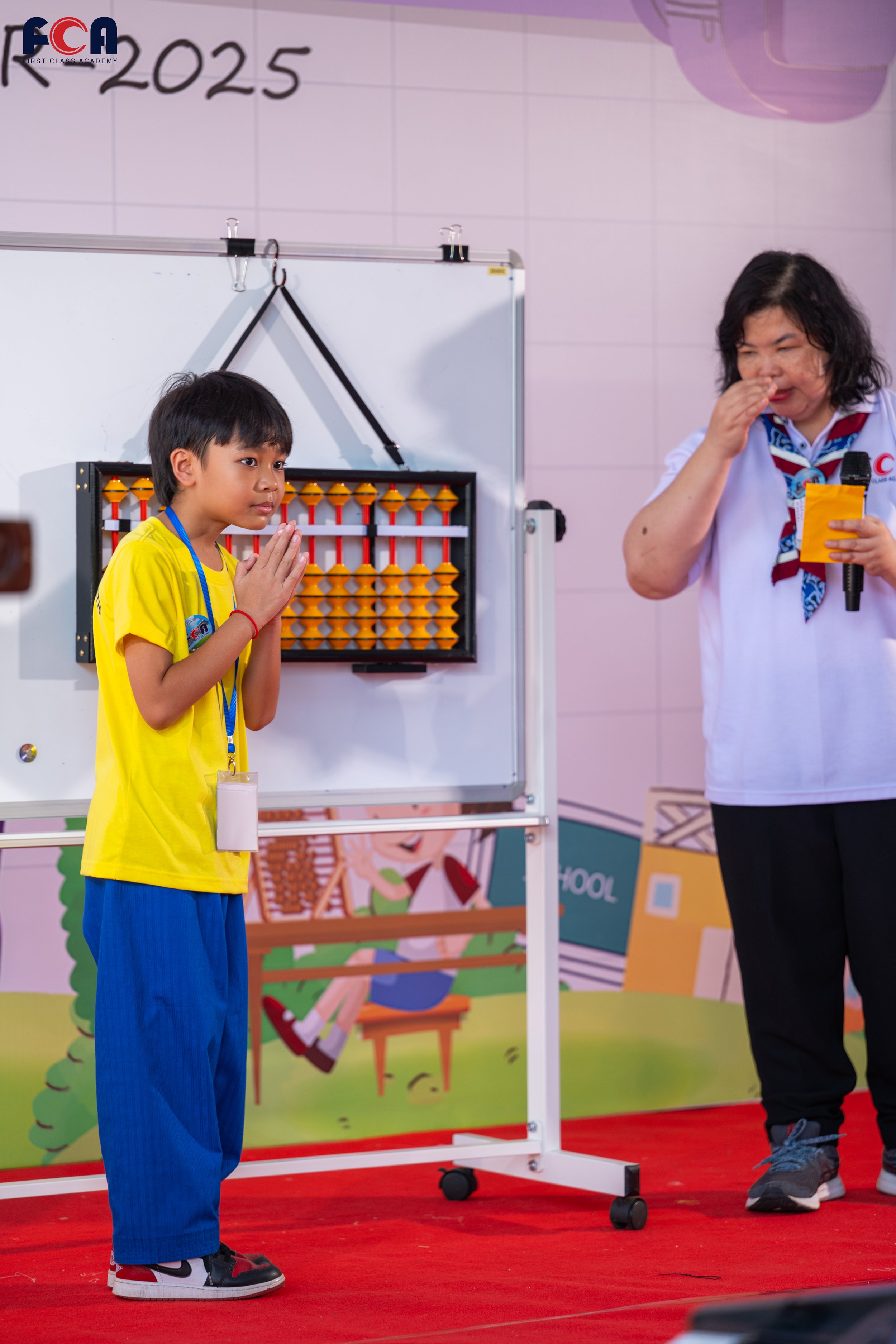A young boy in a yellow shirt and blue pants stands with hands together in a prayer gesture in front of an abacus or counting frame. An adult woman in a white shirt and black pants stands nearby, holding a microphone and touching her nose. They are in a classroom or presentation setting with a whiteboard and colorful backdrop featuring cartoon children and school-themed illustrations.