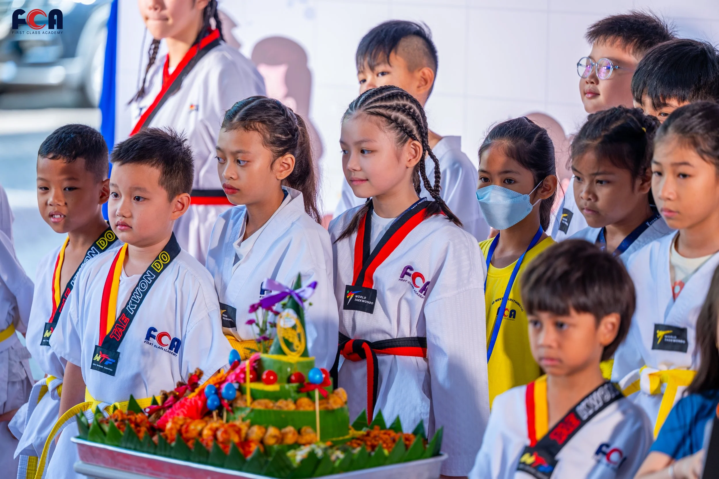 Group of children in martial arts uniforms gathered around a colorful, decorated cake during a celebration or event.