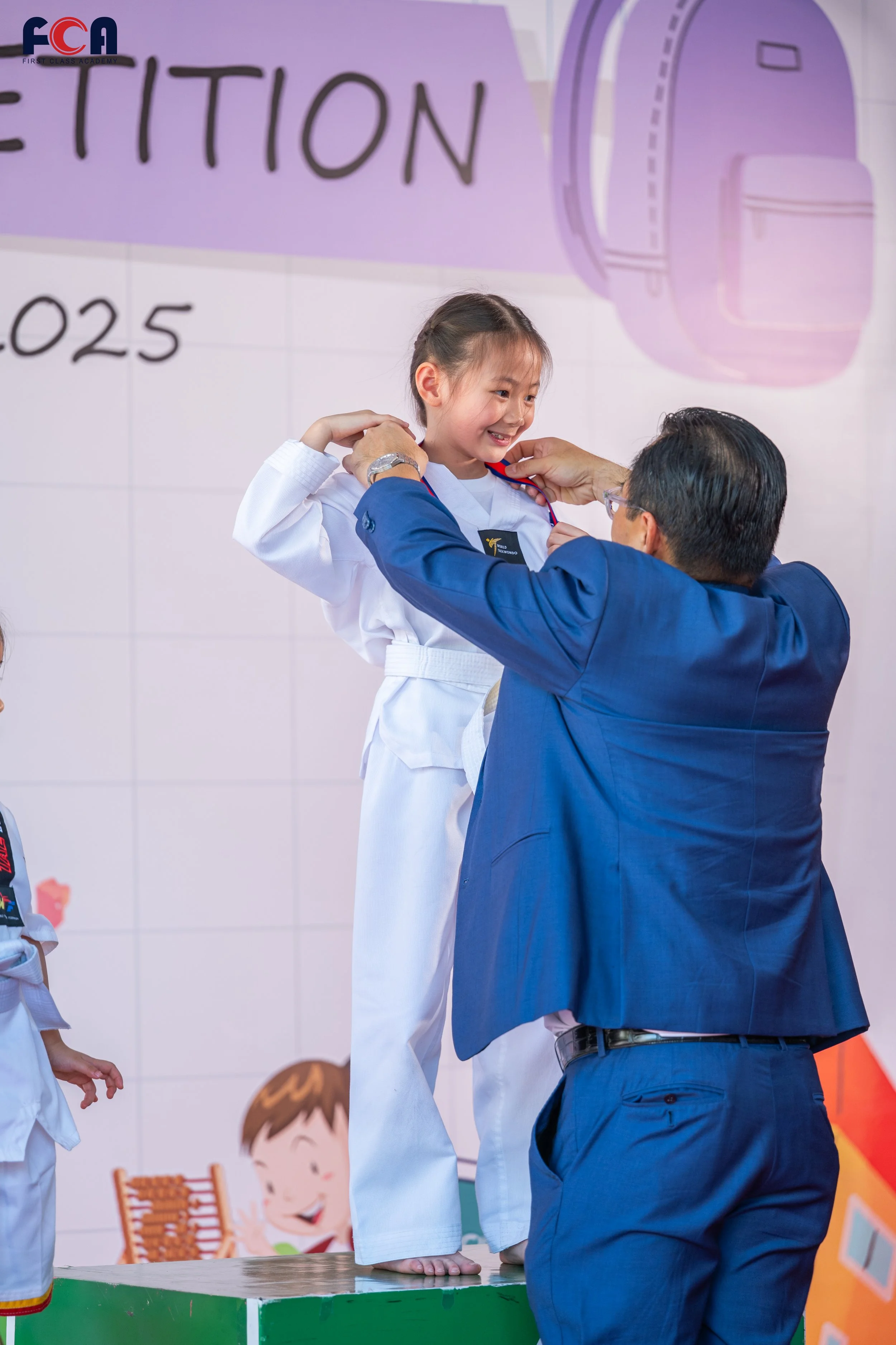 A young girl in a martial arts uniform receiving a medal from a man in a blue suit during an award ceremony, with a backdrop featuring words and a large illustration of a backpack.