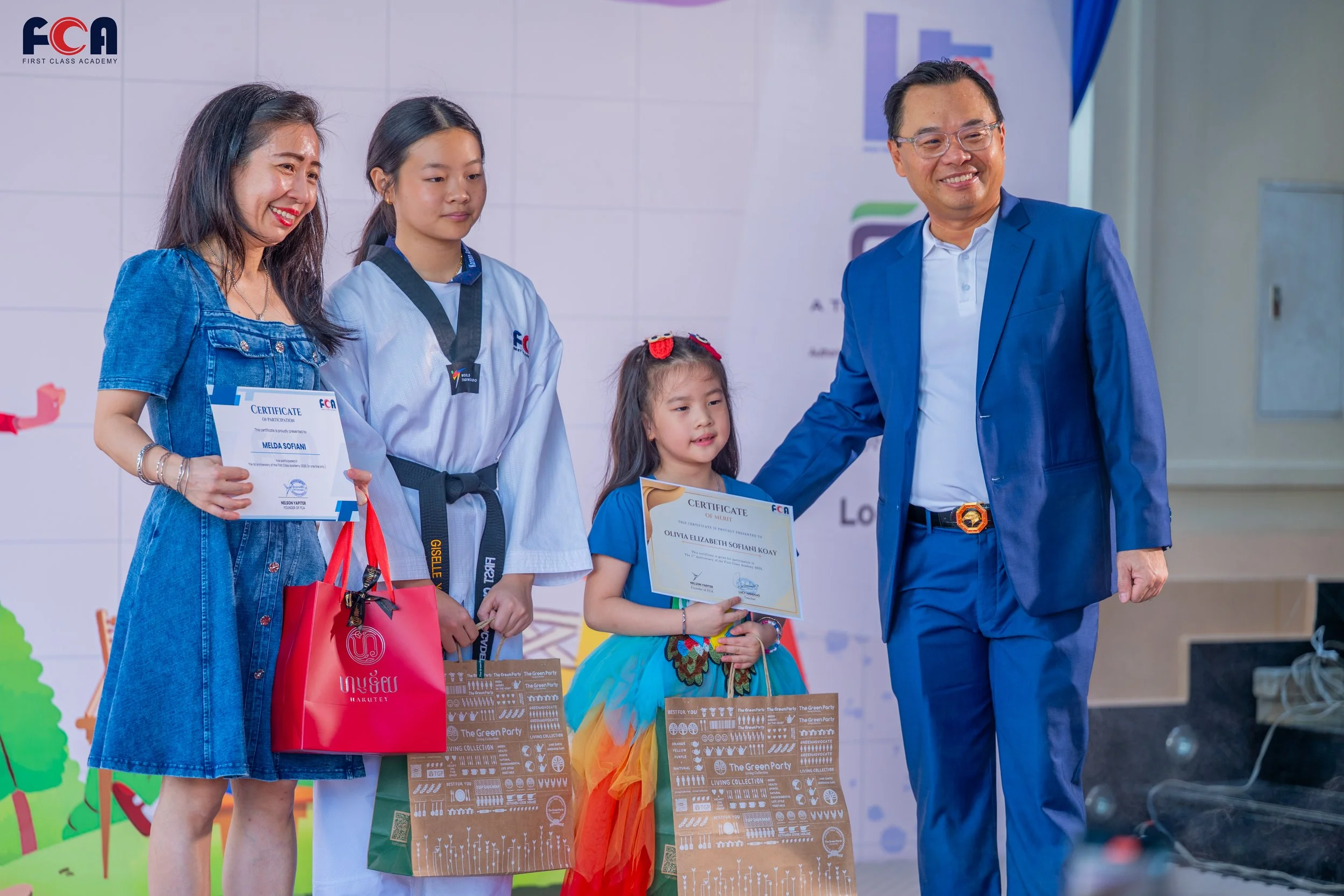 A group of four people, three girls and one man, standing on a stage during an award ceremony. The girls are holding certificates and gift bags, and the man is smiling and placing his hand on one girl's shoulder. There is a backdrop with the logo of a martial arts academy.