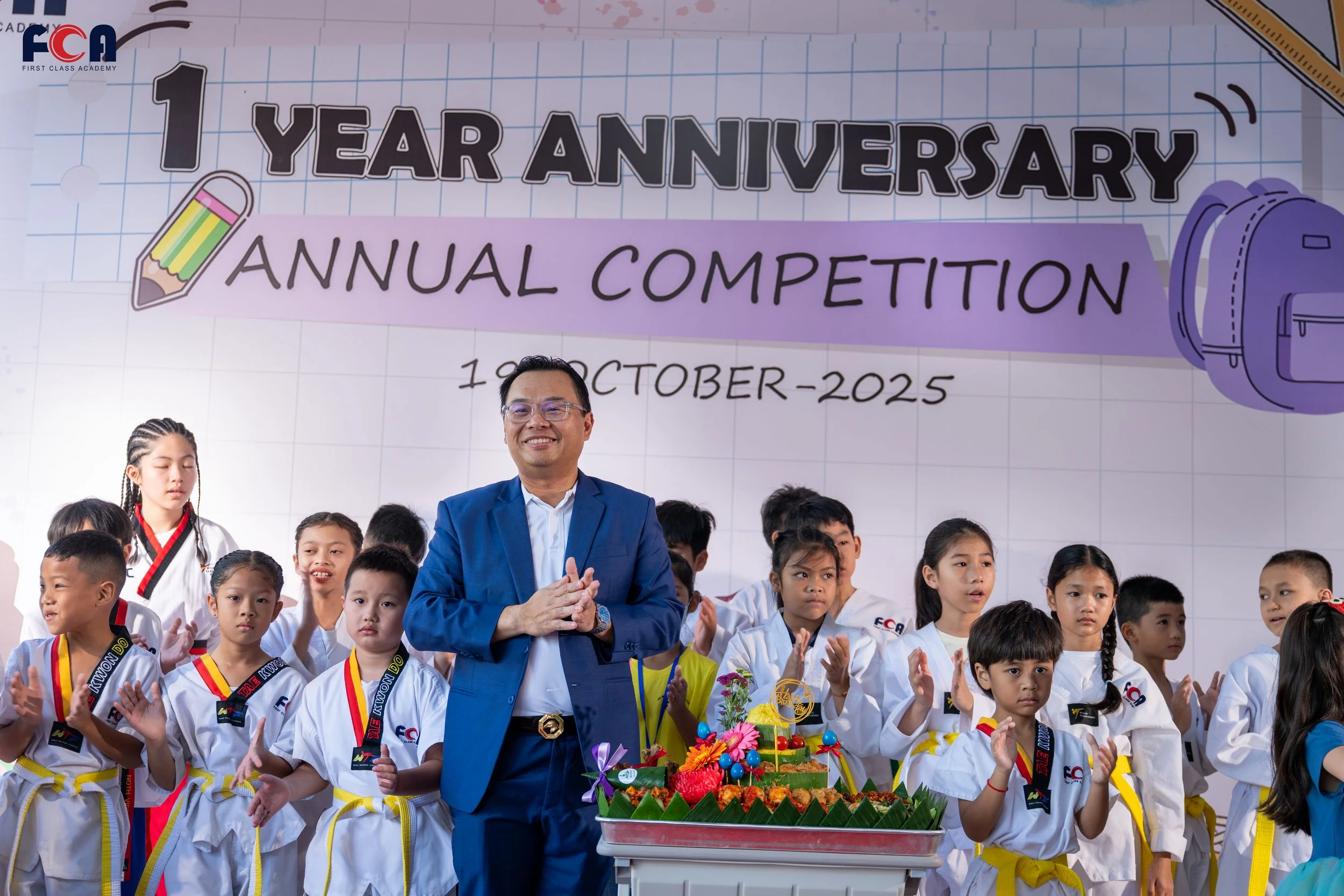 A group of children and a man in a blue suit celebrating a one year anniversary with a traditional Asian cake decorated with flowers and fruits, in front of a backdrop that reads '1 Year Anniversary Annual Competition, October 2025', with cartoon school supplies illustrations.