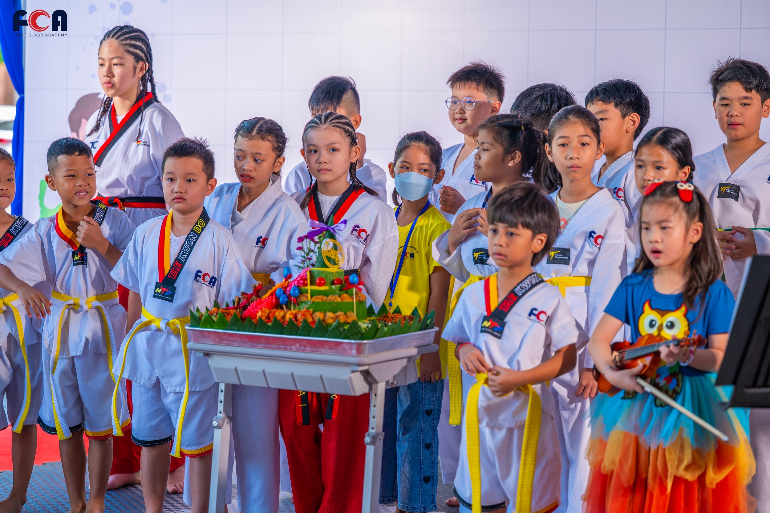 Children dressed in martial arts uniforms gather around a colorful birthday cake with a cake topper and fruit decoration, with some playing a guitar, in a martial arts academy setting.