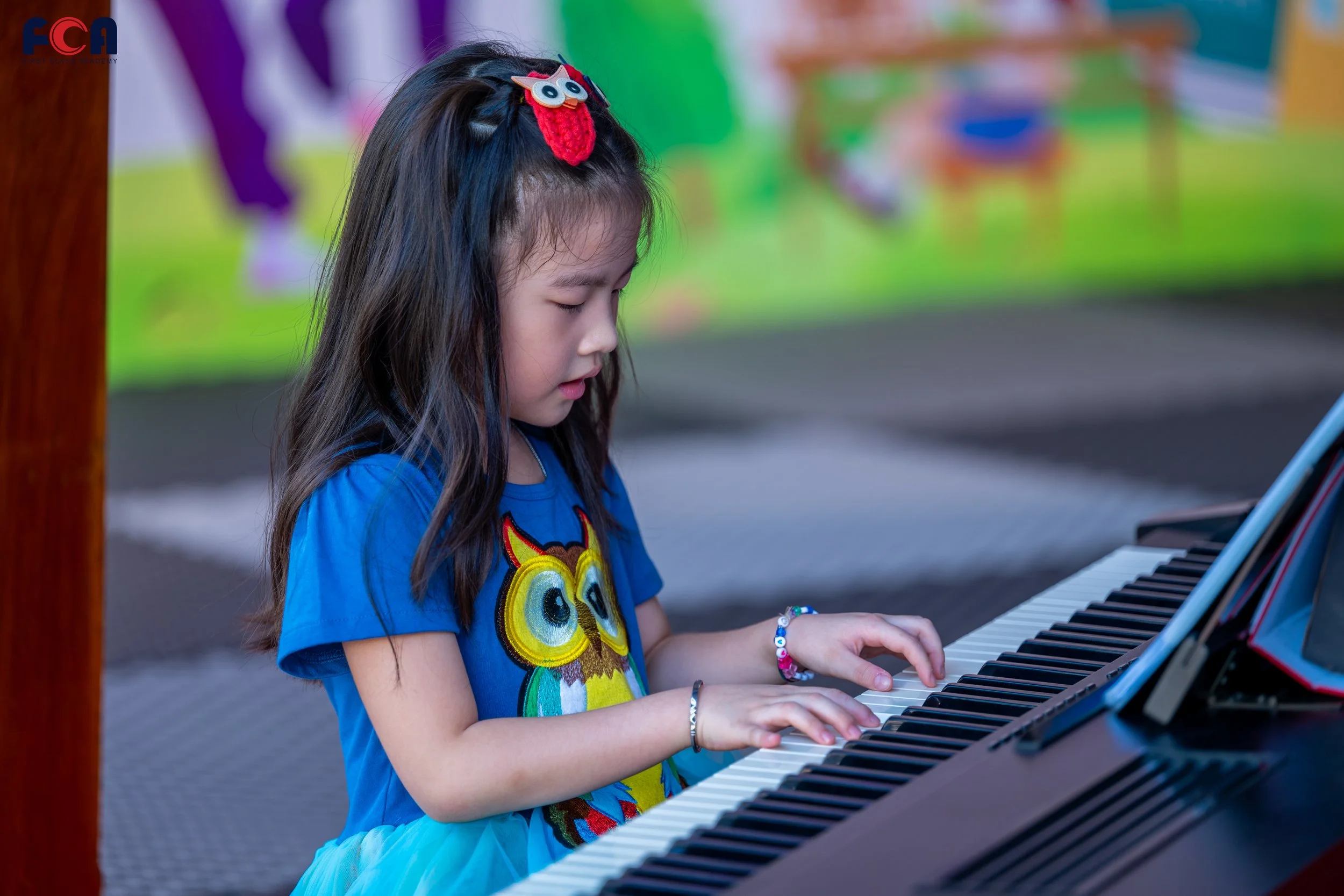 A young girl with long dark hair, wearing a blue owl graphic t-shirt and a teal skirt, is playing a black digital piano outdoors. She has a red owl hair clip and colorful bracelets, with her eyes closed and her hands on the piano keys.