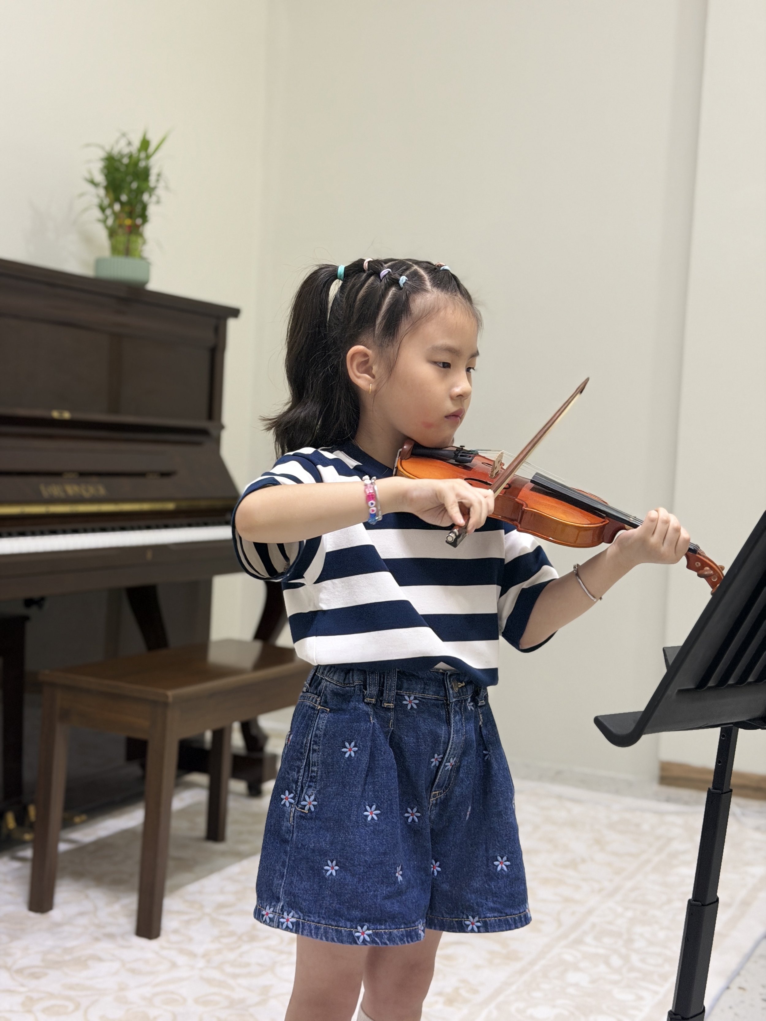 A young girl playing the violin in a room with a piano and a music stand.