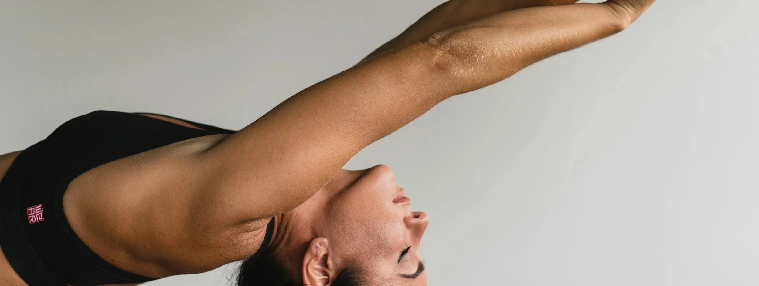 A woman doing a yoga pose with her arms raised overhead, wearing a black sports bra, against a light gray background.