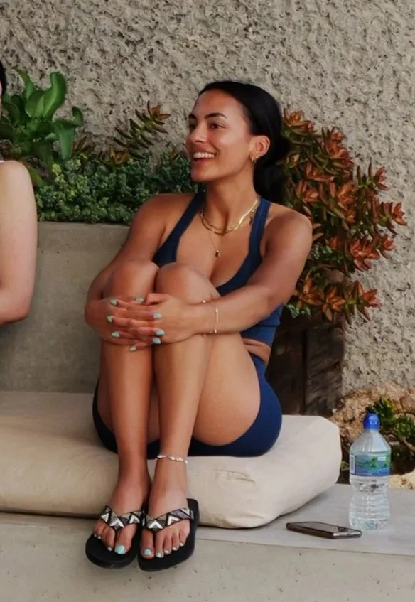 A woman sitting on an outdoor bench with her arms wrapped around her knees, smiling. She has dark hair, is wearing a navy blue tank top and shorts, and black sandals with white geometric patterns. There is a water bottle and a smartphone on the bench next to her, with green and reddish plants in the background.