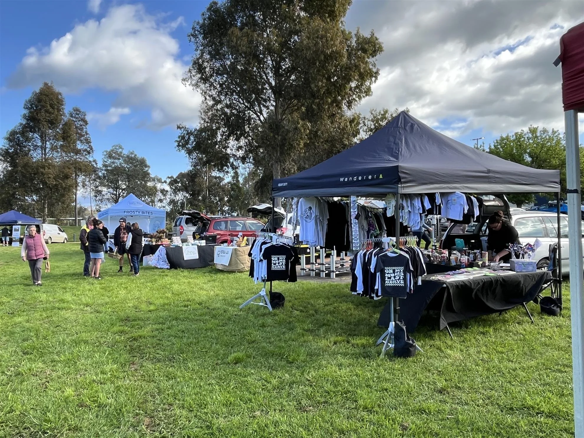 Outdoor market scene with tents and vendors selling clothing and other items on a grassy field, with people browsing.