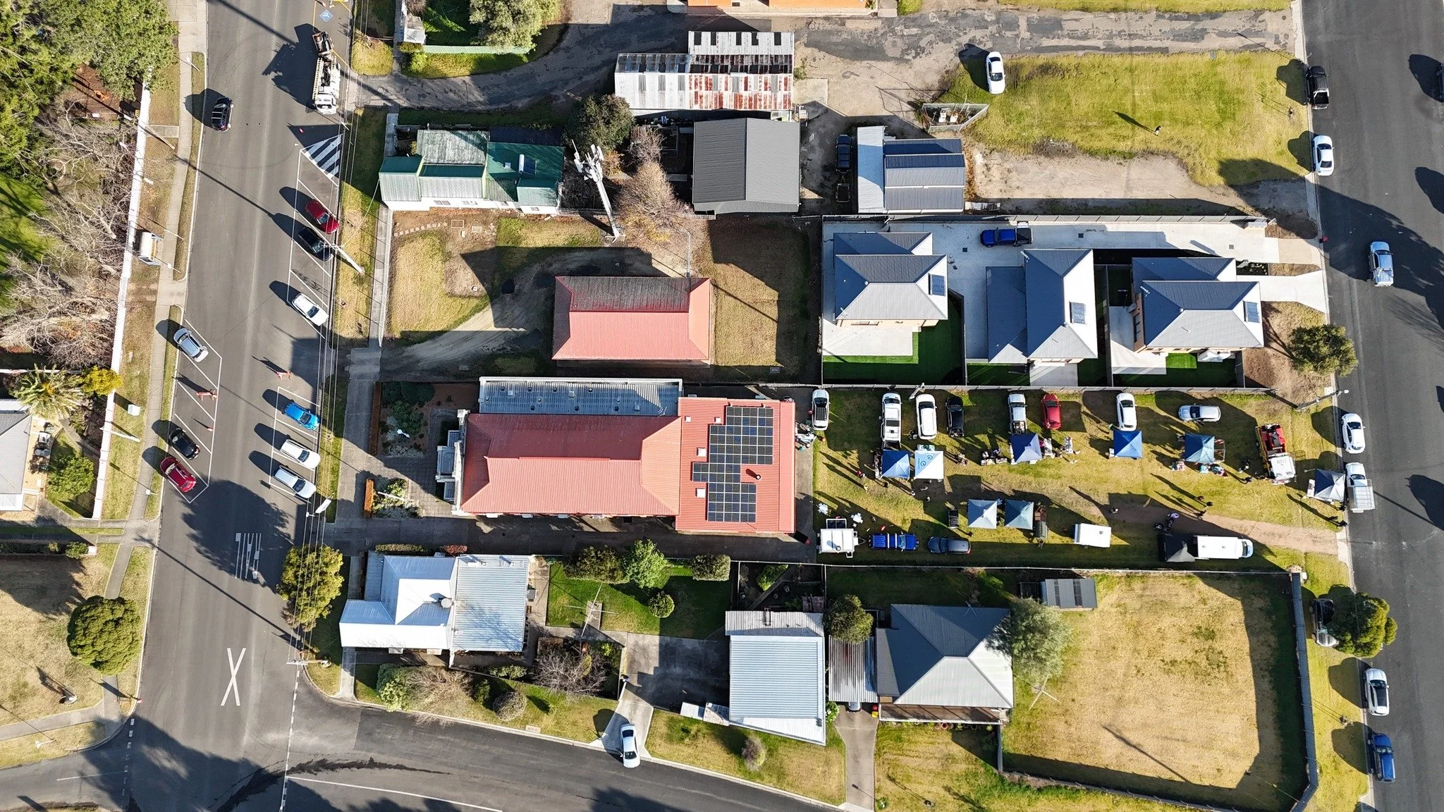 Aerial view of residential houses, parked cars, and a community event with tents on a sunny day.