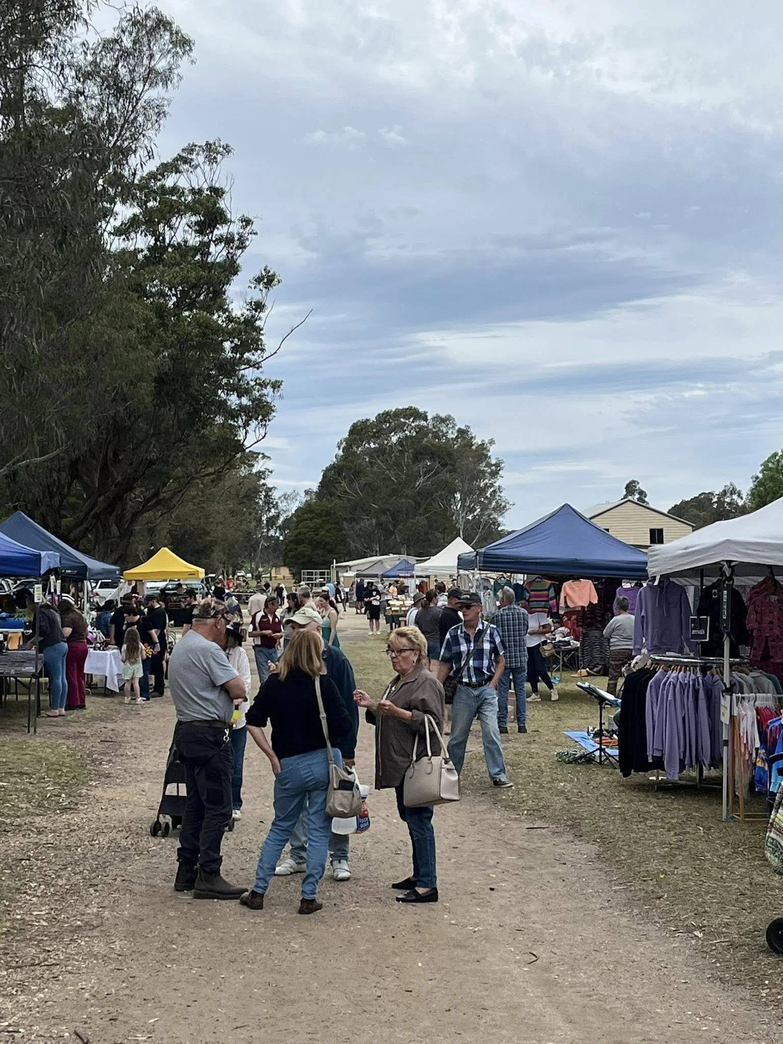 People attending an outdoor market with vendor tents, browsing stalls, and socializing on a dirt pathway under a cloudy sky.