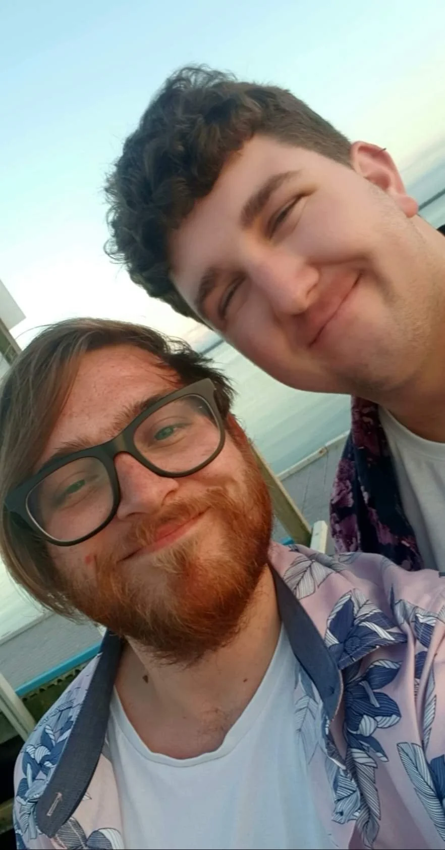 Two young men smiling and taking a selfie outdoors, with a beach and ocean in the background.