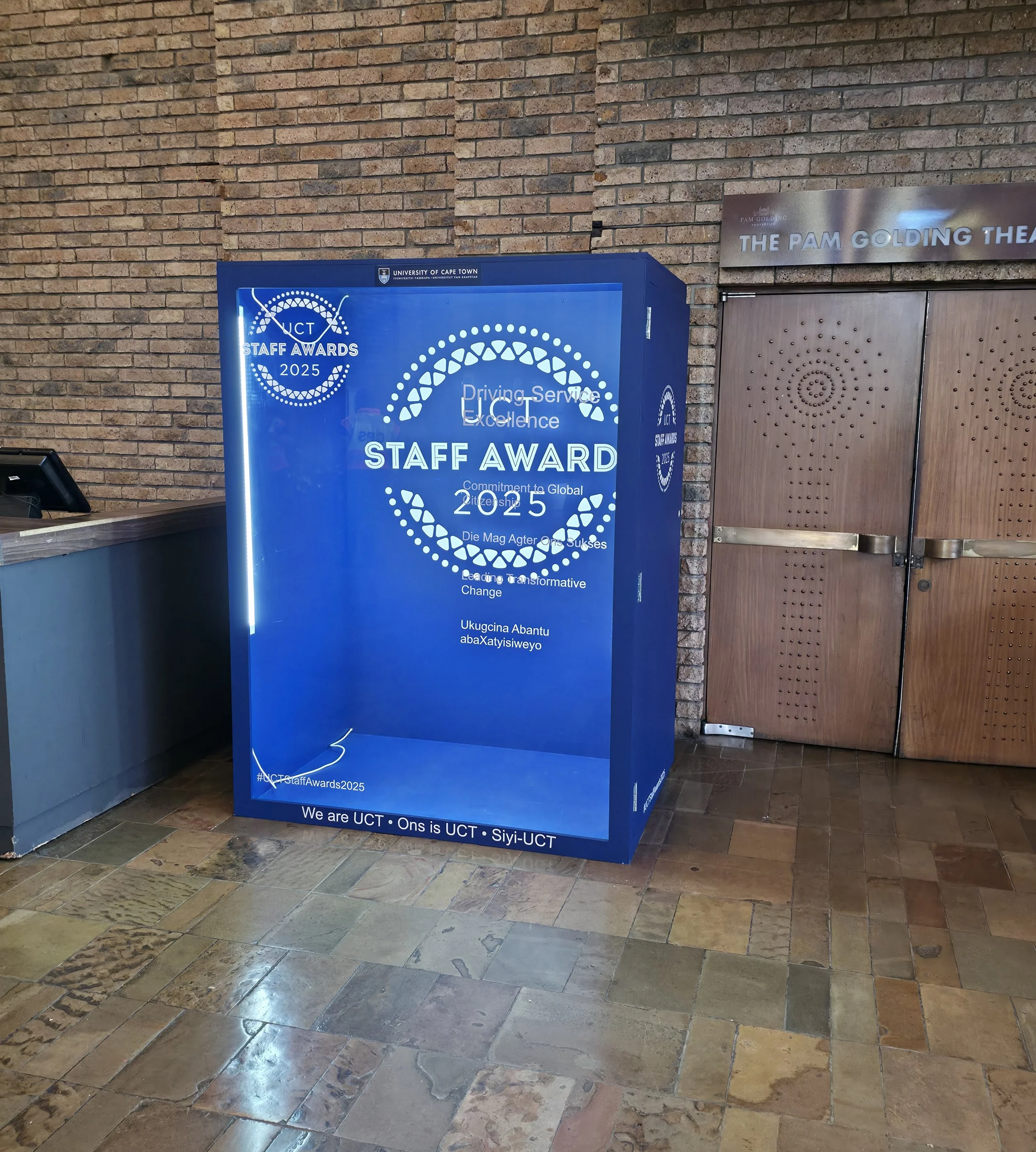 A blue digital display board with text about the University of Cape Town Staff Awards 2025, placed against a brick wall, next to a wooden door.