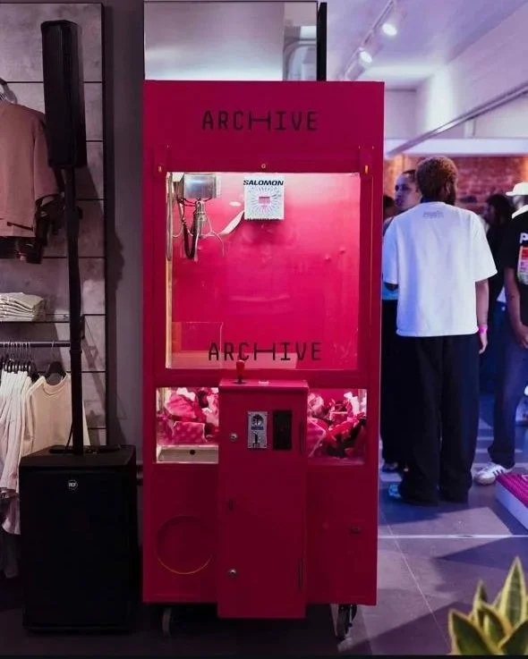 A pink claw machine game labeled 'ARCHIVE' with stuffed animals inside, located in an indoor retail space with people in the background.