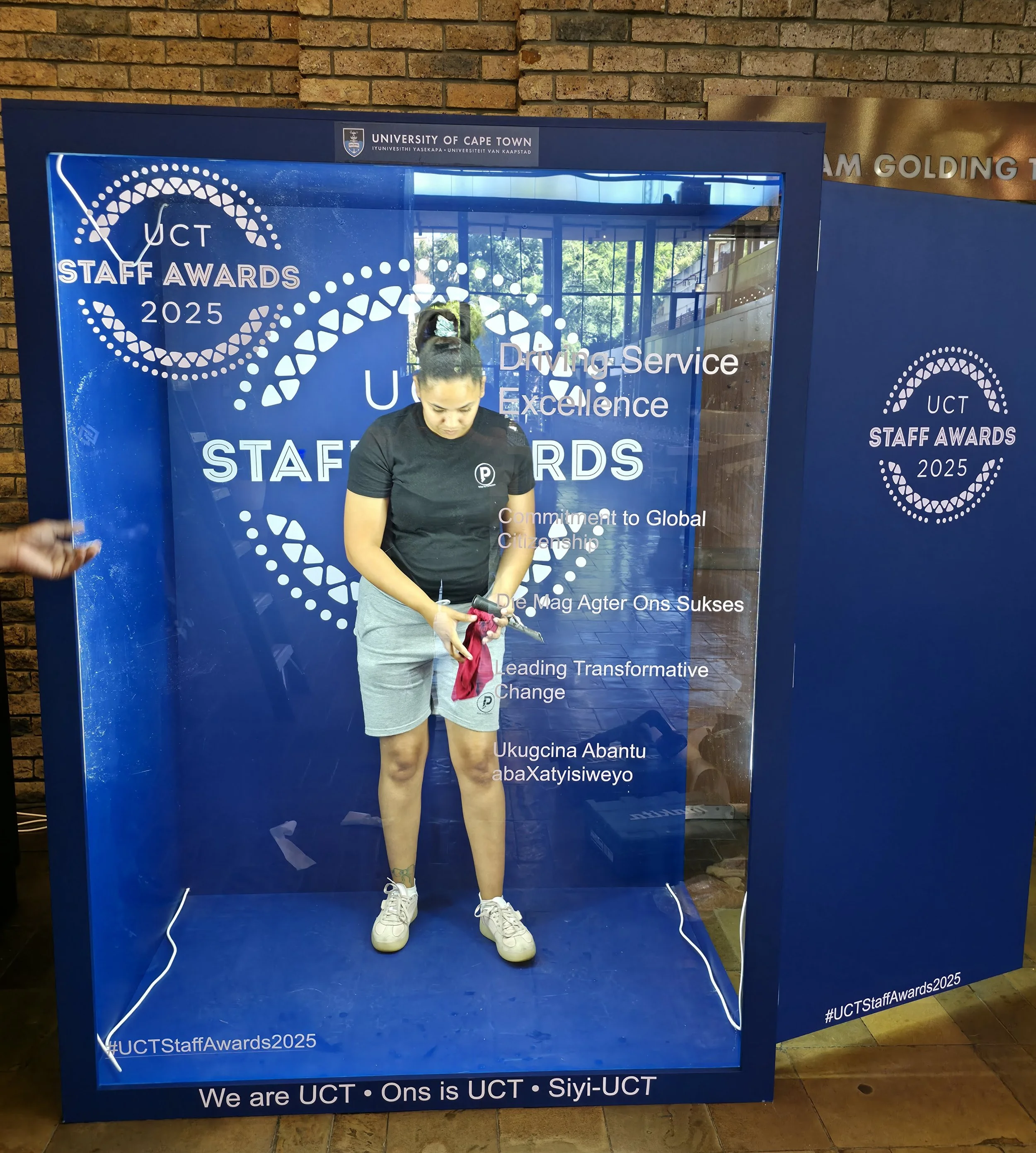 Young woman with a ponytail wearing a black T-shirt, gray shorts, and white sneakers, standing inside a glass frame with blue background and text at the UCT Staff Awards 2025 event, holding a pink cloth and a tool.