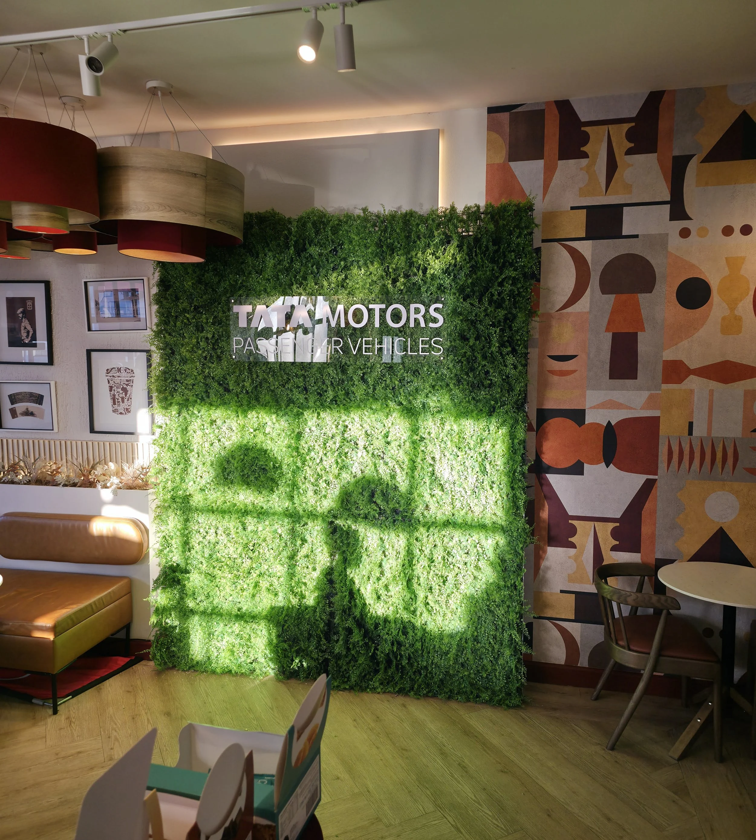 Interior of a room with a green living wall displaying the words "TATA MOTORS PASSENGER VEHICLES" illuminated in white. The room has modern decor including framed pictures, a brown cushioned bench, a small round table with two chairs, and colorful ab