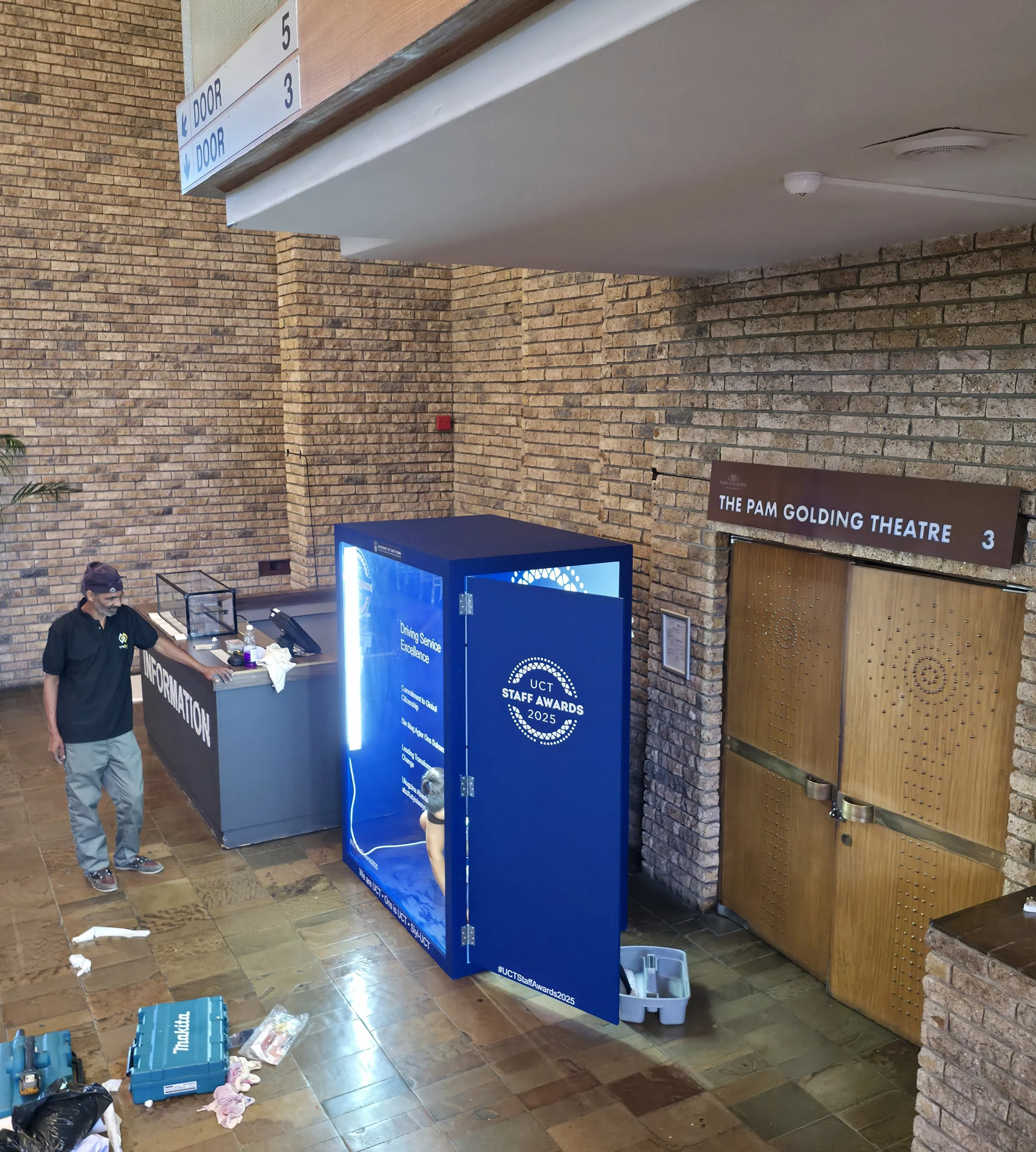 Reception area at The Pam Golding Theatre with a man near an information desk, a bright blue digital display booth for UCT Staff Awards 2025, and wooden double doors. The scene shows a tiled floor, brick walls, and directional signs for doors.