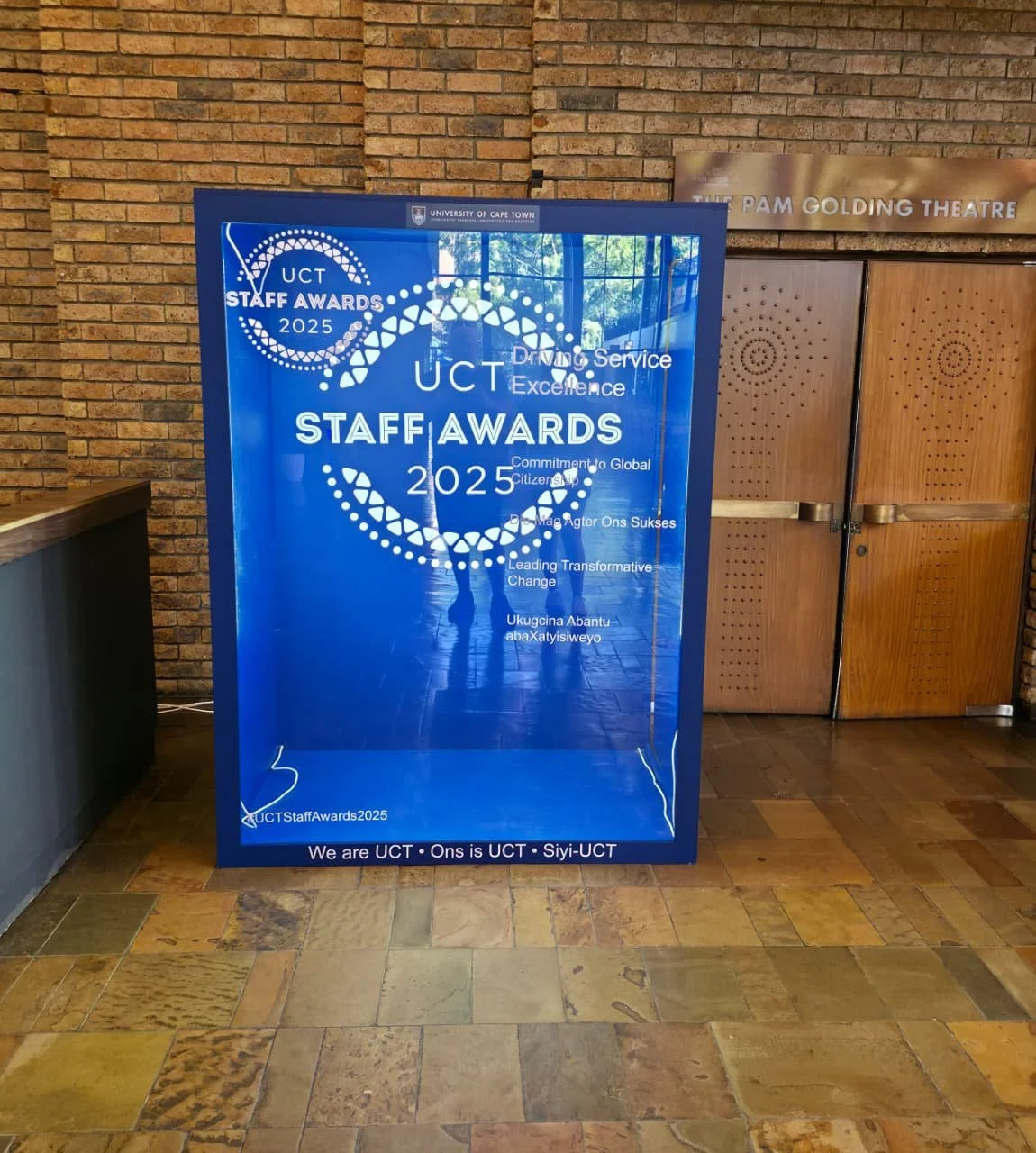 Blue poster announcing the University of Cape Town (UCT) Staff Awards 2025, with details about excellence, global commitment, transformational change, and celebratory quotes, placed in front of a brick wall and wooden door.