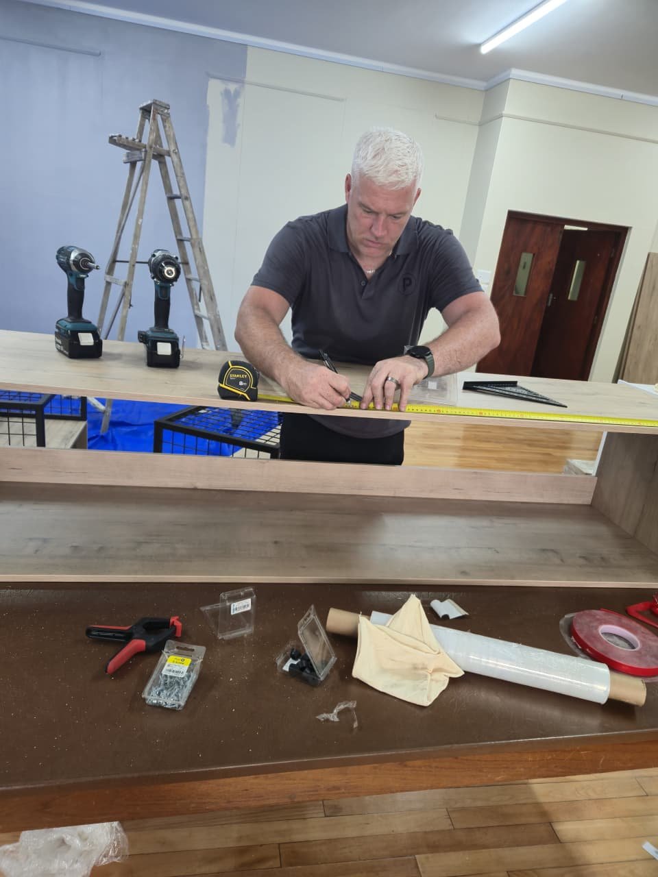 A man with white hair measures and marks a wooden board during a woodworking project, with tools and power drills on the table in front of him.